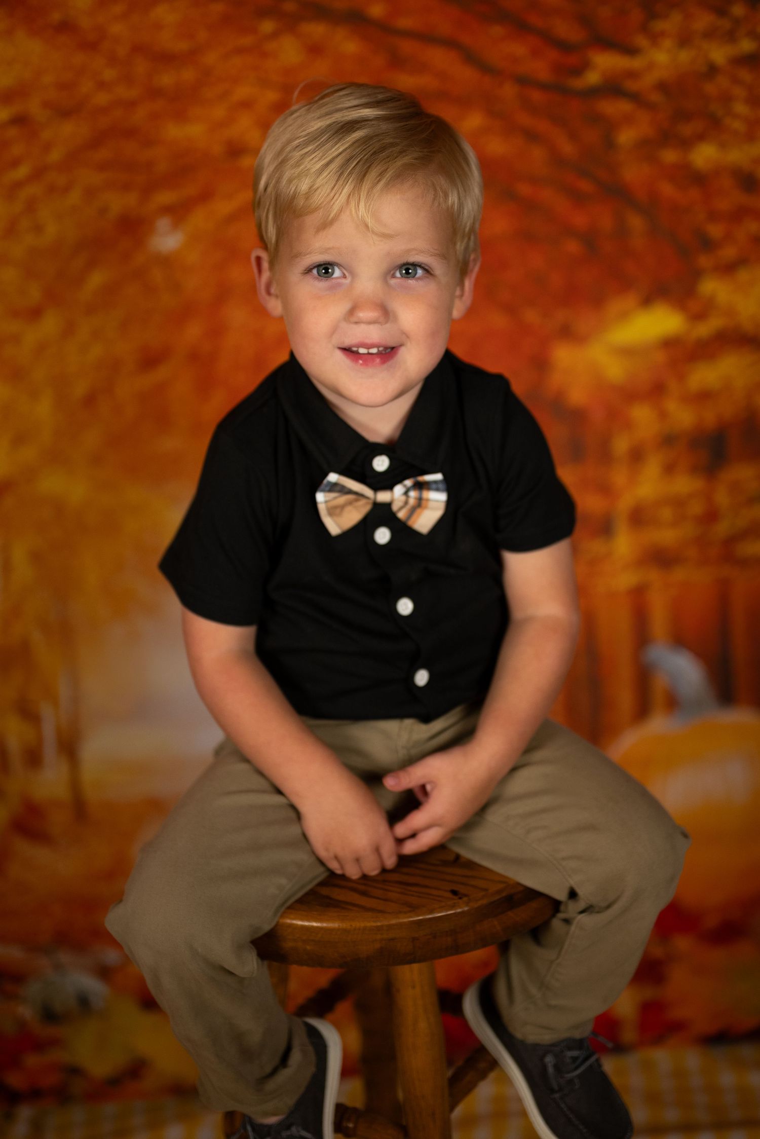 A young child in a black shirt with bow tie sits on a wooden stool against a fall-themed backdrop.