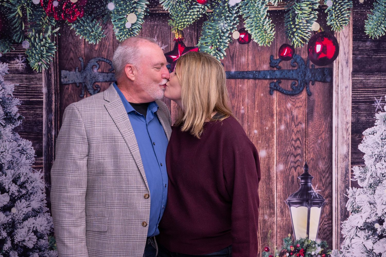 A couple in formal attire shares a tender moment under Christmas decorations and evergreen branches.