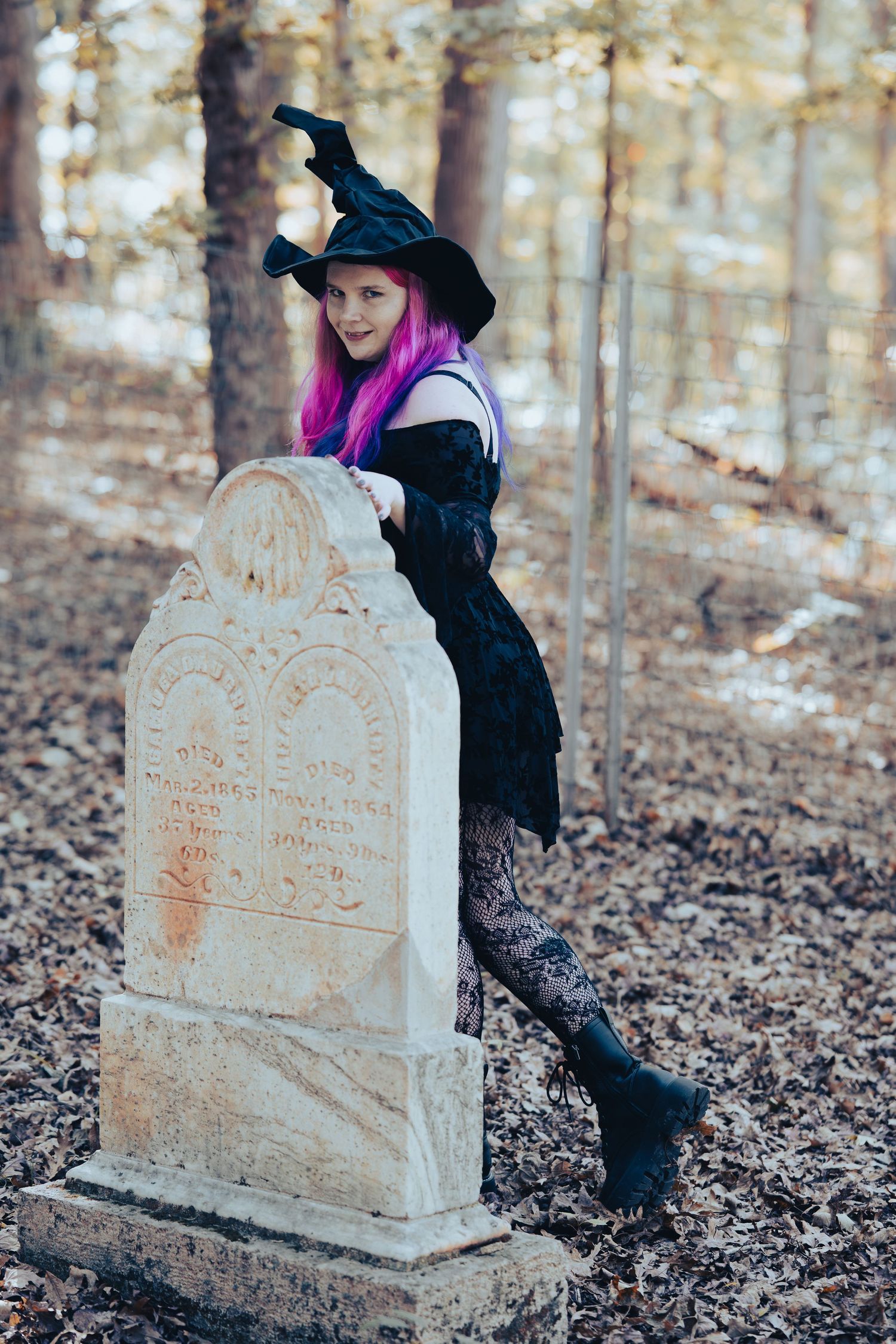 A witch with bright pink hair and black dress poses next to an old weathered gravestone in an autumn forest.
