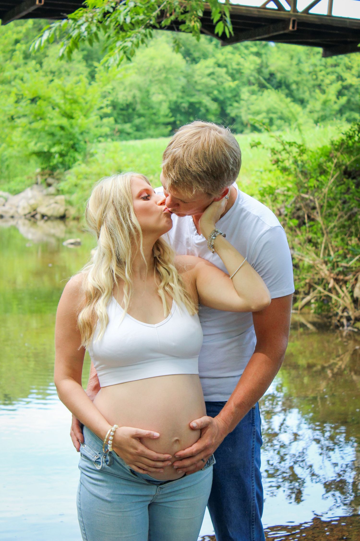 A tender maternity photo captured by a serene pond with lush greenery in the background.