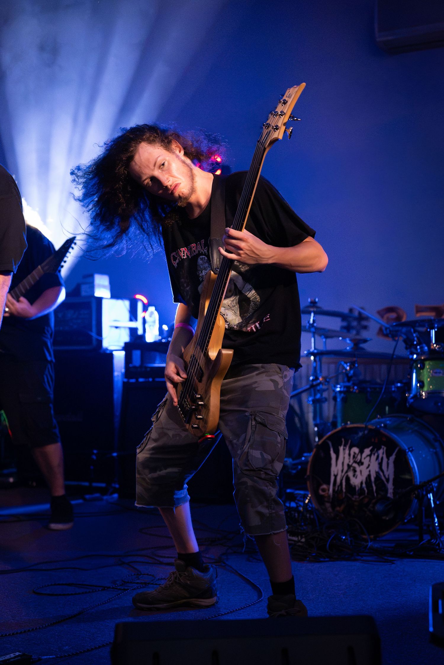 Young male guitarist with long curly hair performs on stage with electric guitar under blue stage lights at a rock concert.