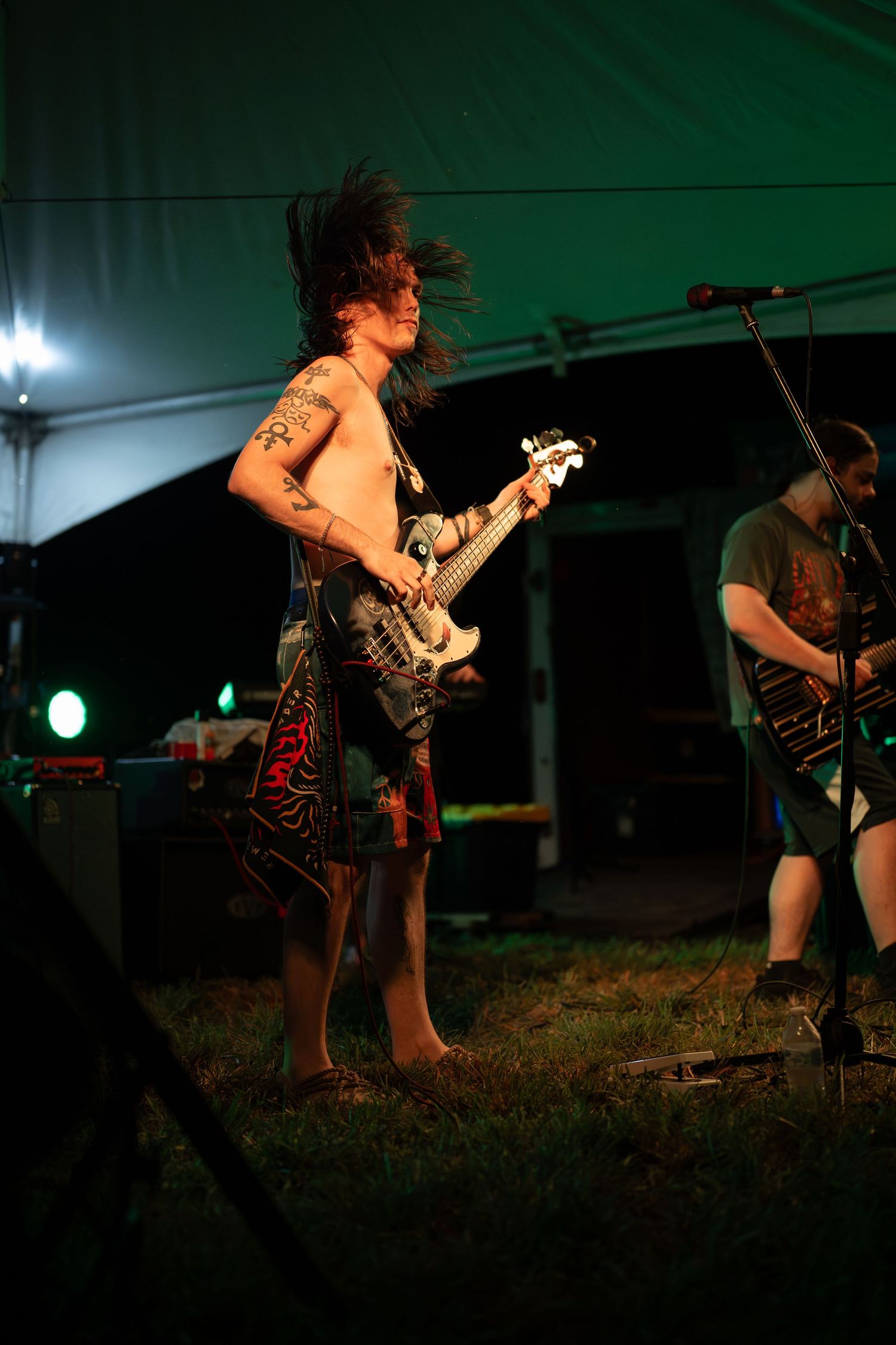 Shirtless tattooed guitarist with mohawk plays electric guitar at outdoor rock concert under green tent lights at night.