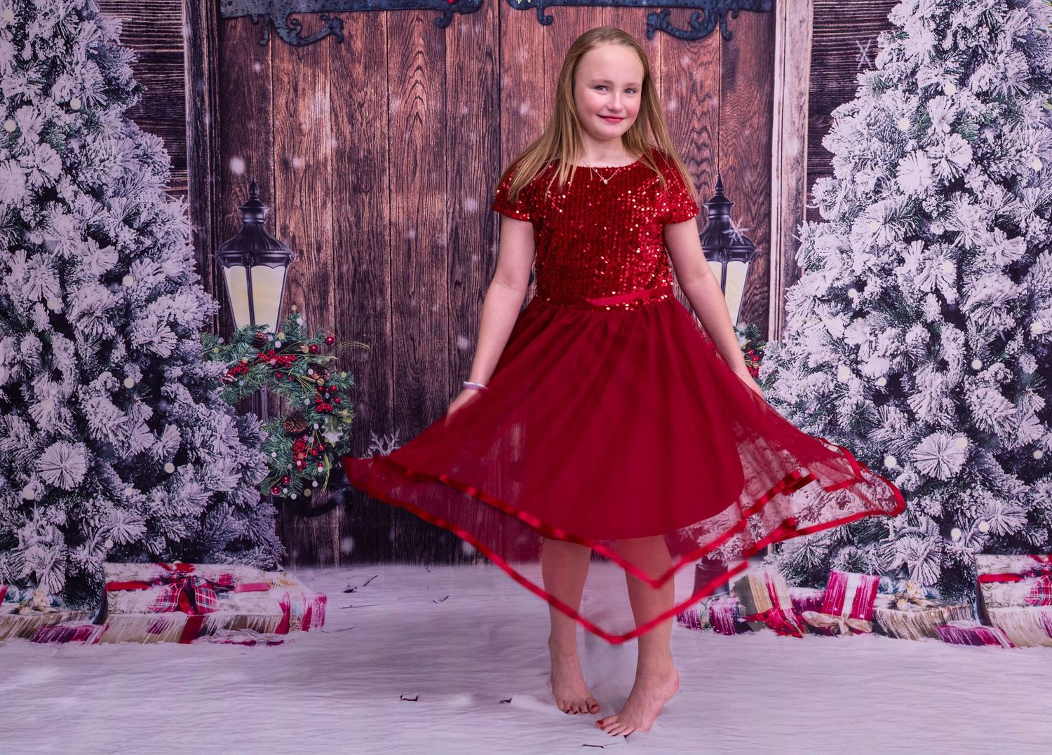 A sparkly red holiday dress twirls against a backdrop of snowy flocked trees and rustic wooden doors.