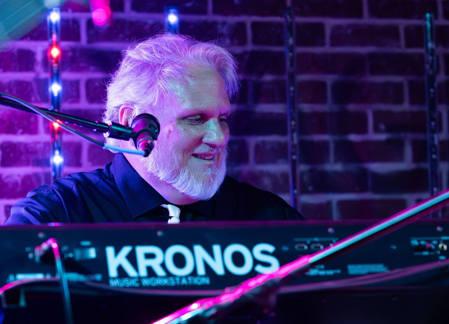 A concert stage illuminated with vibrant purple and blue lighting features a keyboard setup with 'KRONOS' branding.