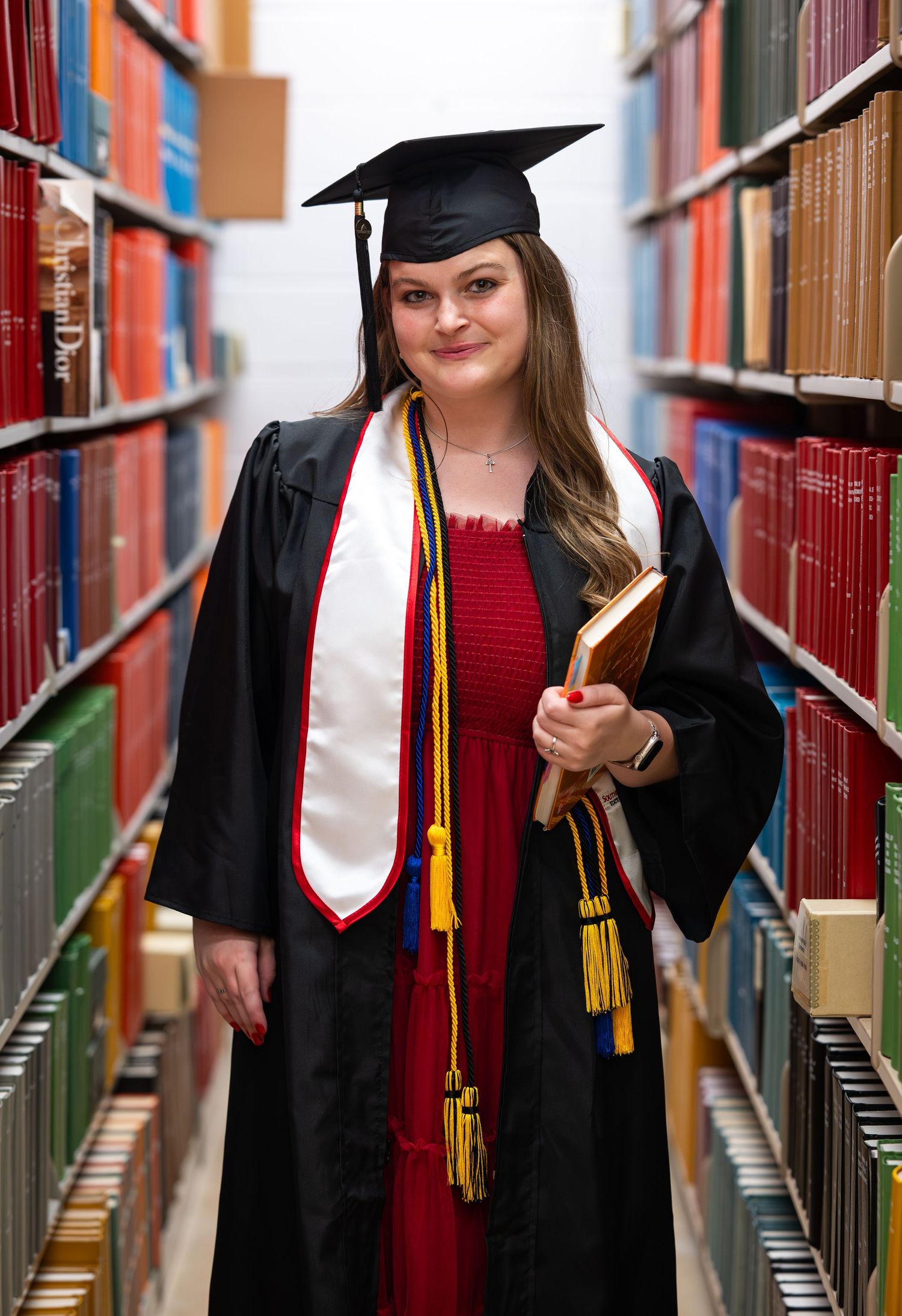 A smiling female graduate in black cap and gown holds books between library shelves, wearing gold tassels and white stole.