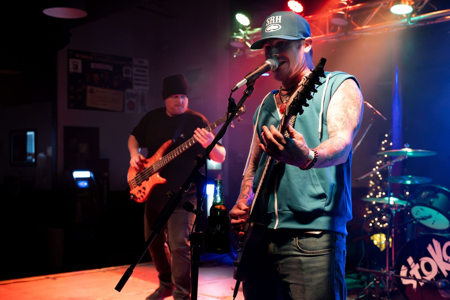 Musicians perform on stage with electric guitars under colorful stage lighting at a live concert venue.