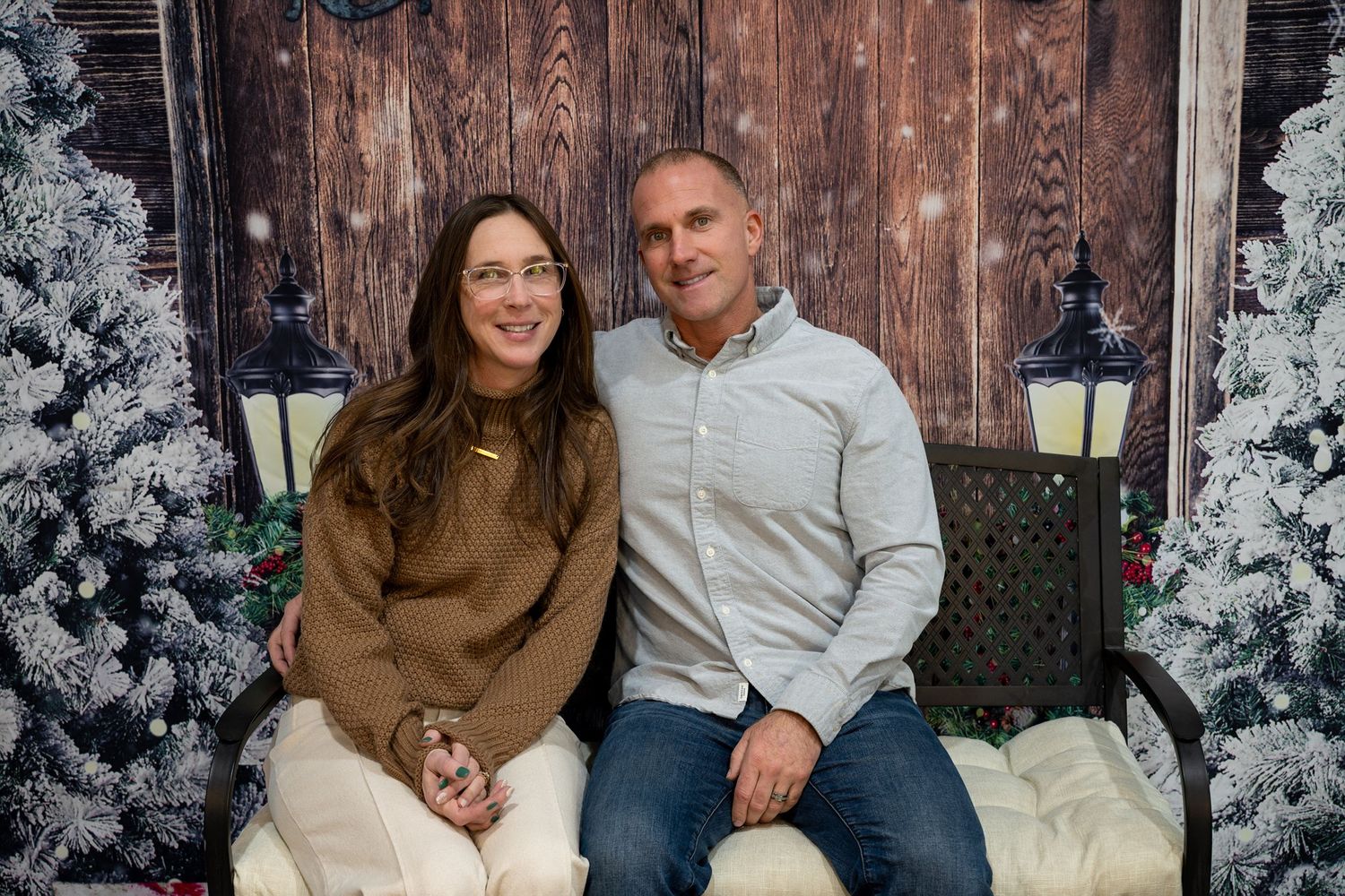 A couple sits together on a bench surrounded by snowy winter decor and frosted Christmas trees in a studio setting.