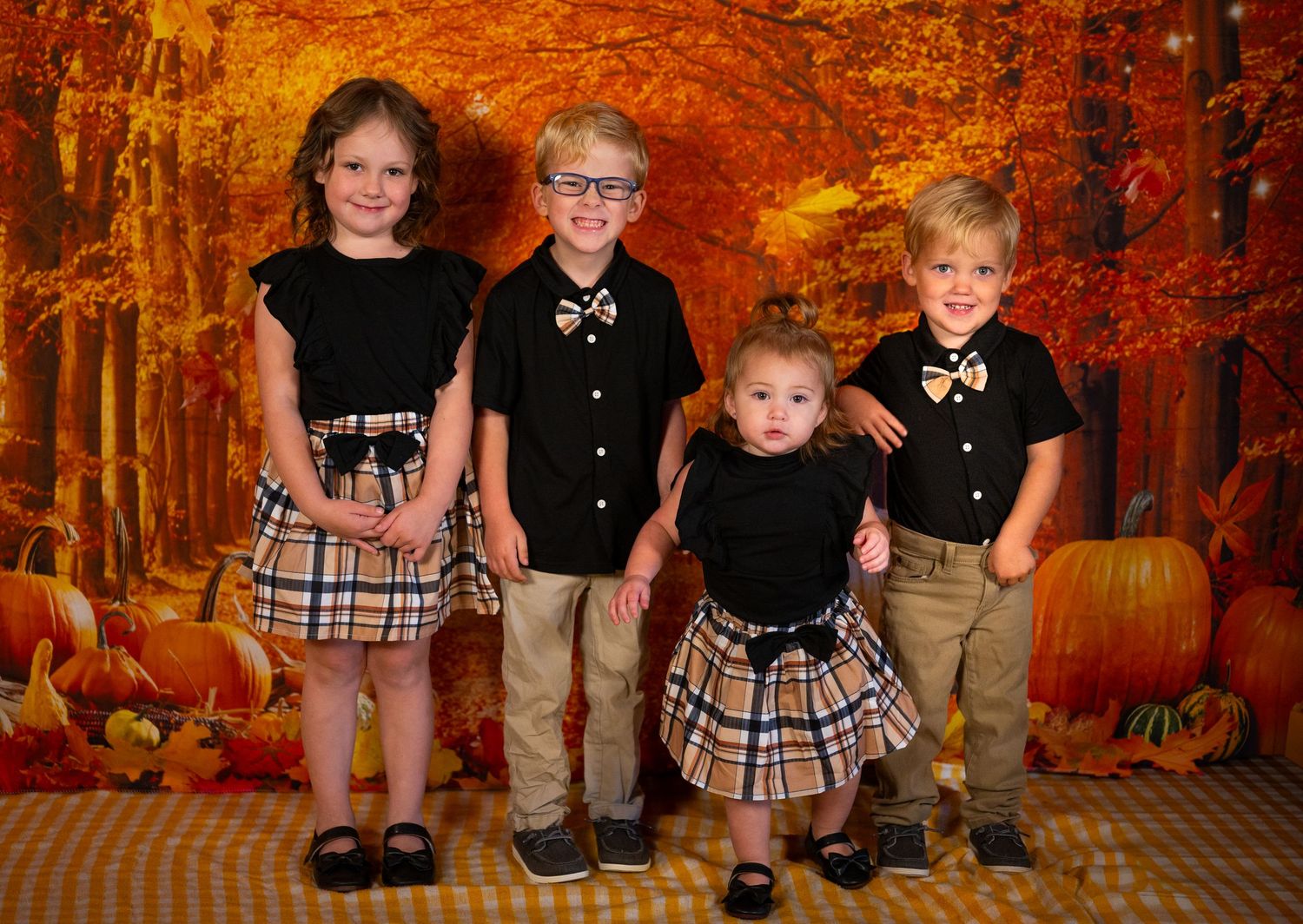 Four children in coordinating black and plaid outfits pose together against an autumn backdrop.