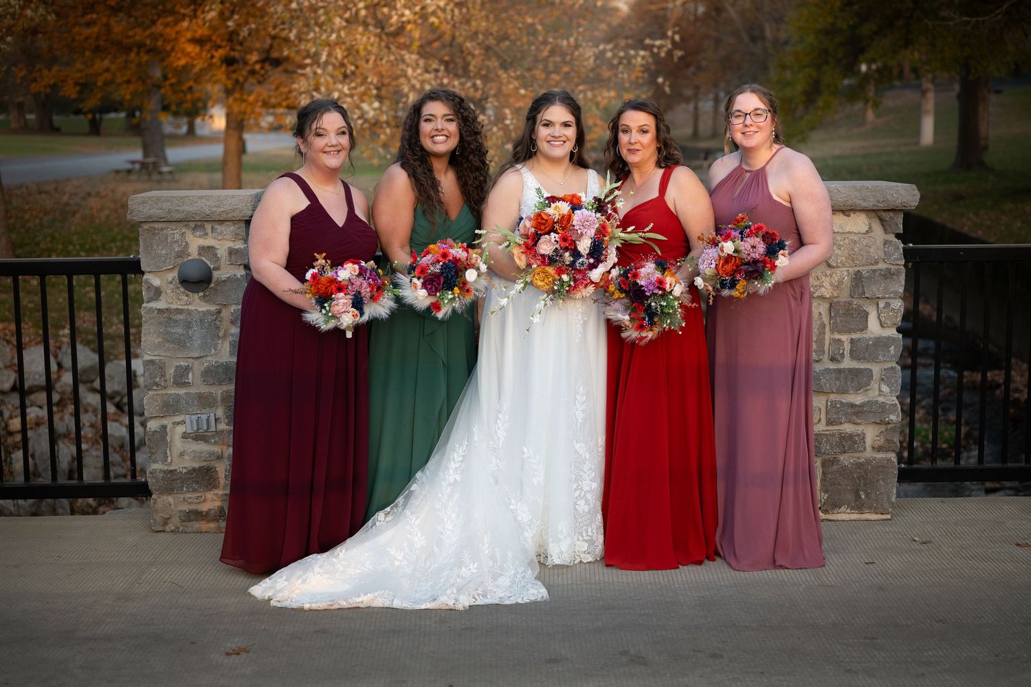 Wedding party in burgundy dresses poses with bride against stone wall backdrop.