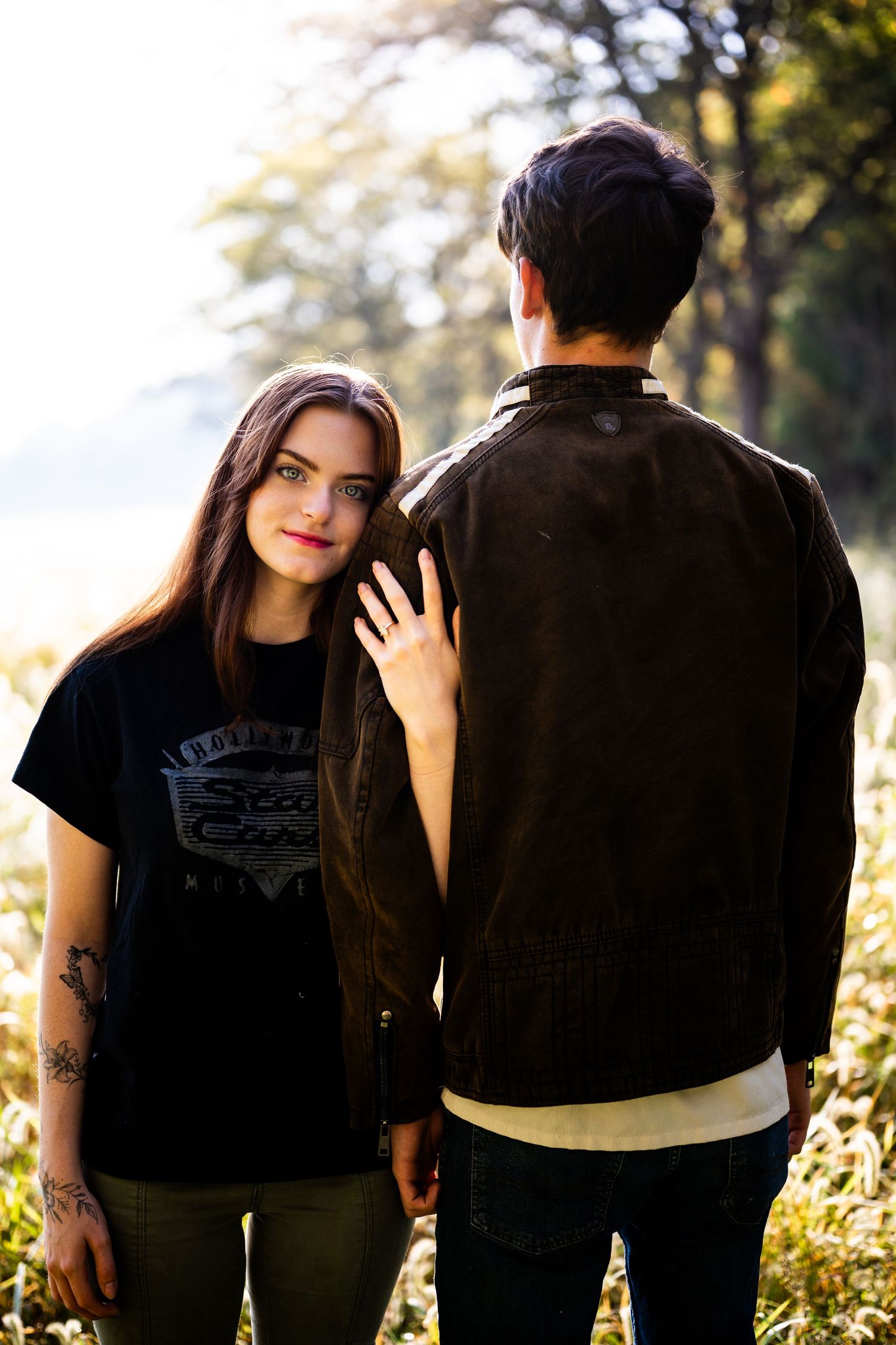 A couple in black clothing embraces while standing in sunlit nature during a romantic photoshoot.
