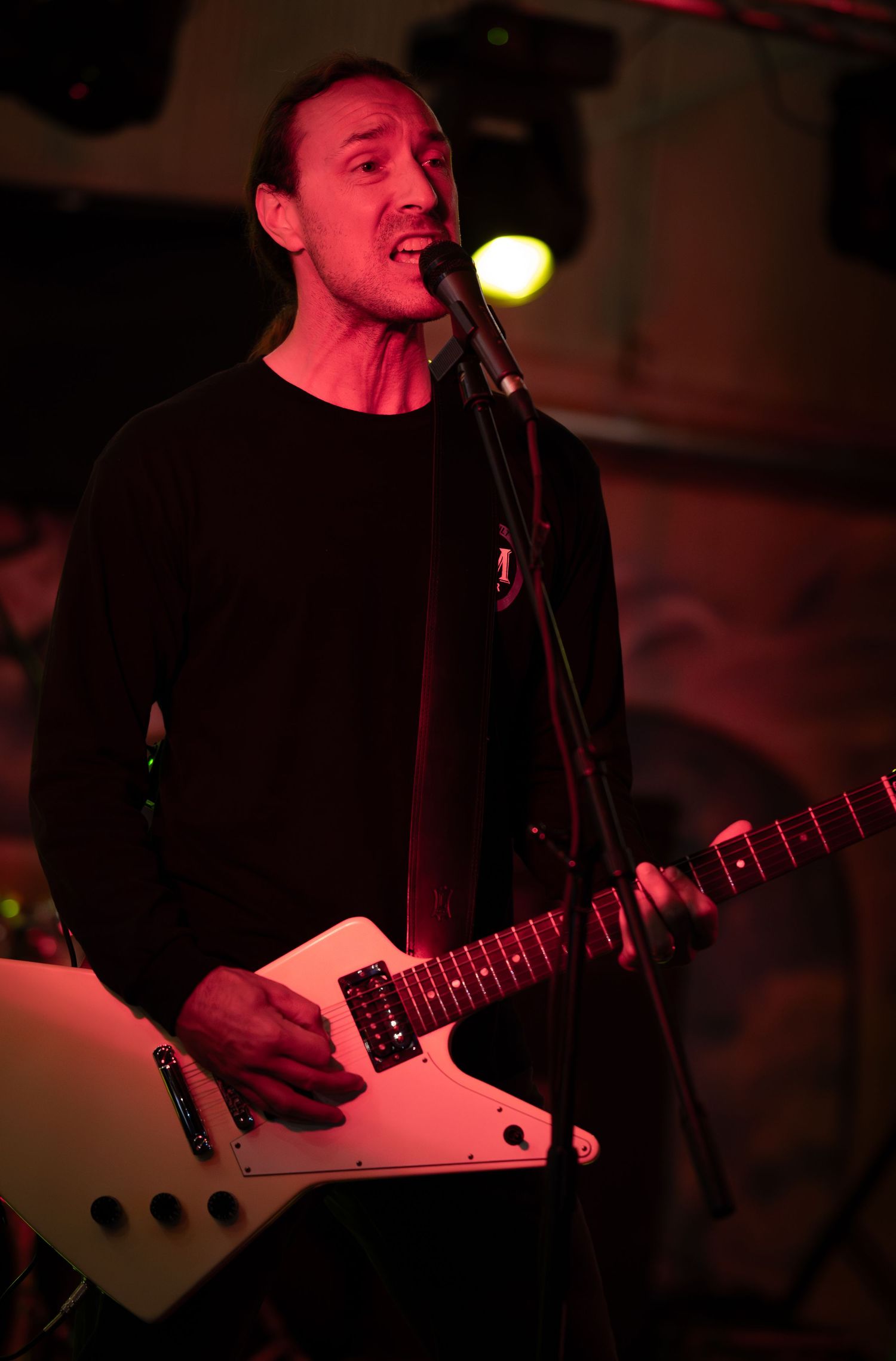 Musician performs on stage with a white electric guitar under dramatic red lighting during a live concert performance.