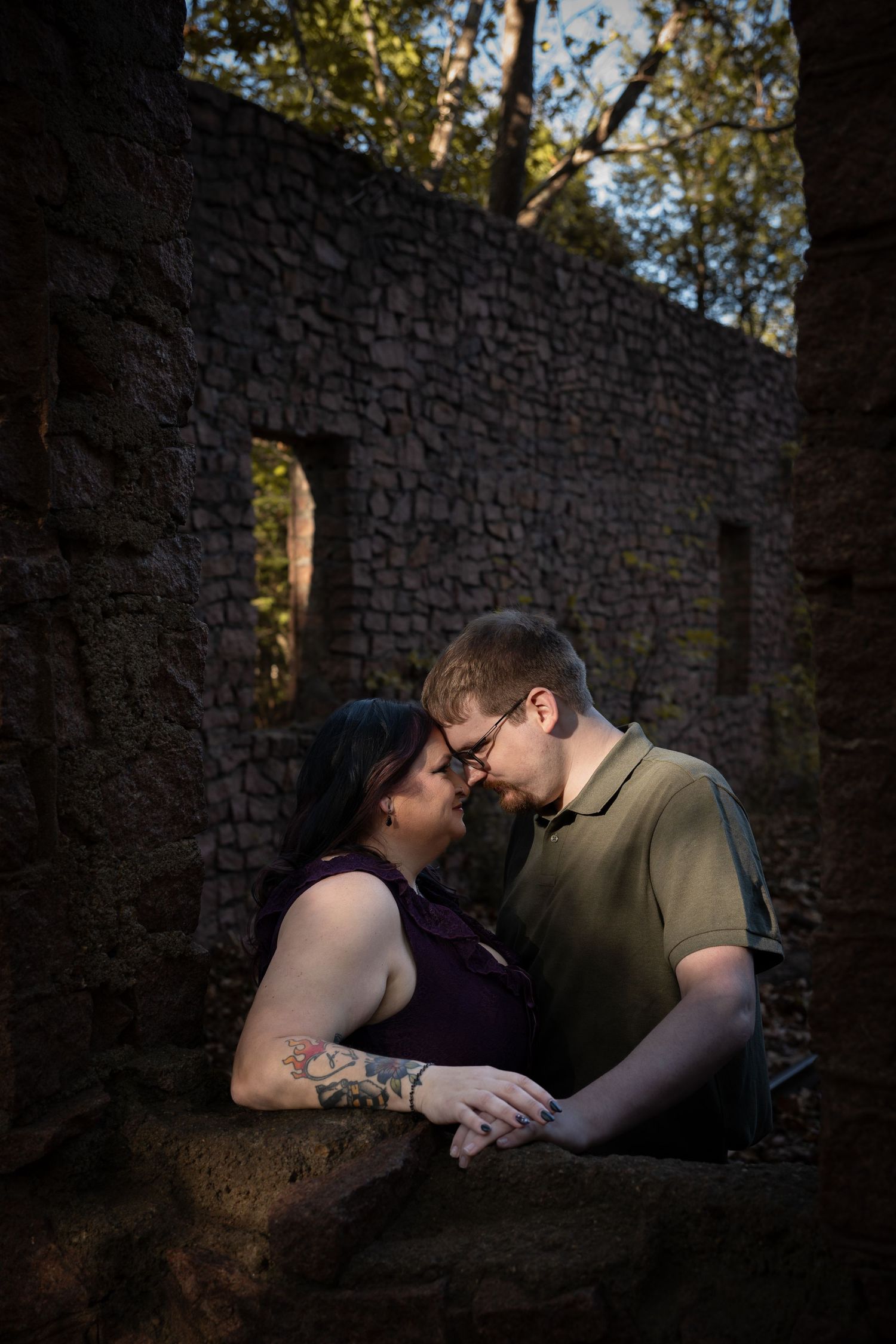 A romantic couple shares an intimate moment against a stone wall backdrop in moody natural lighting.