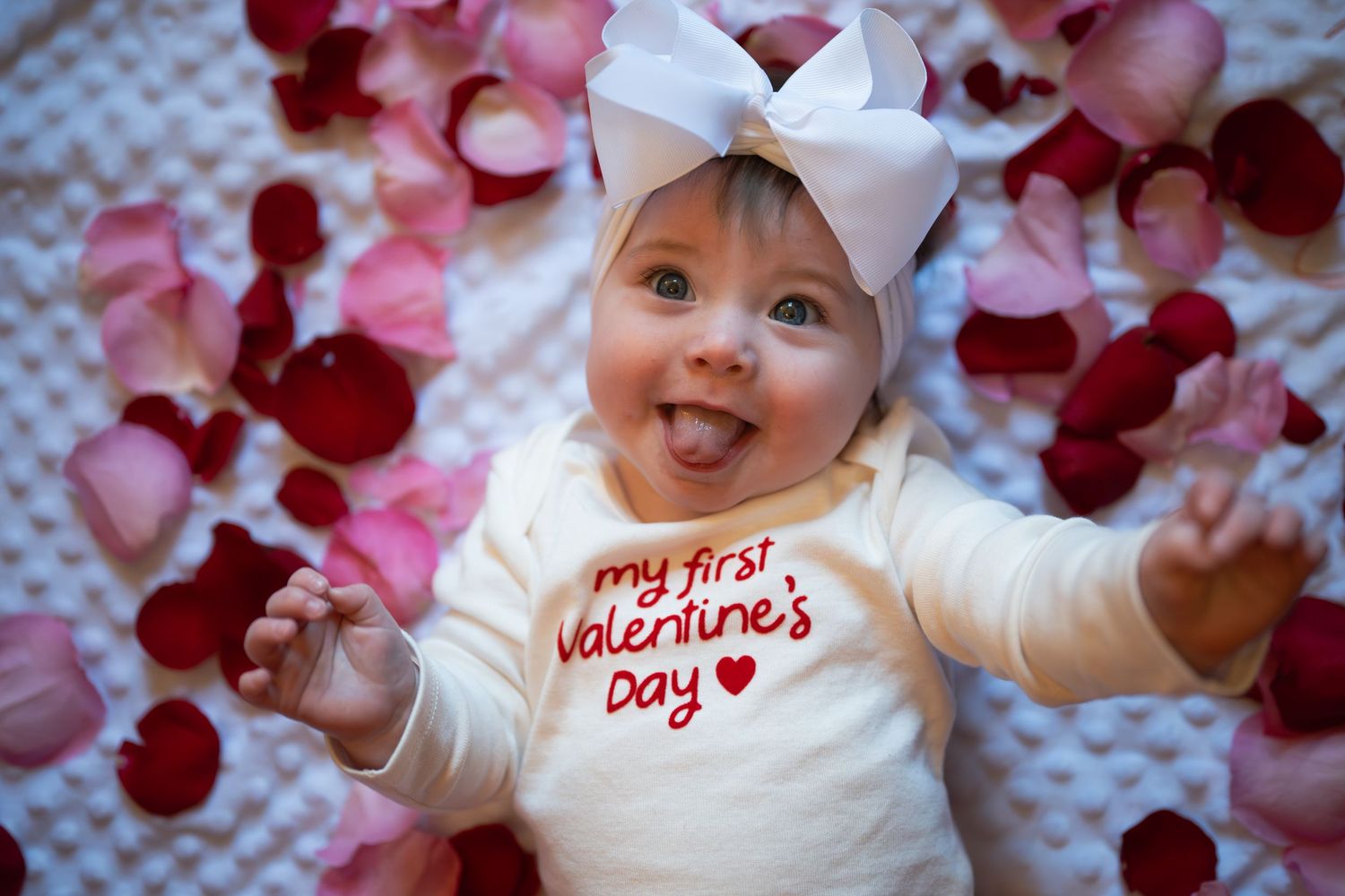 A baby in white clothing lies on pink and red rose petals with a white bow headband.