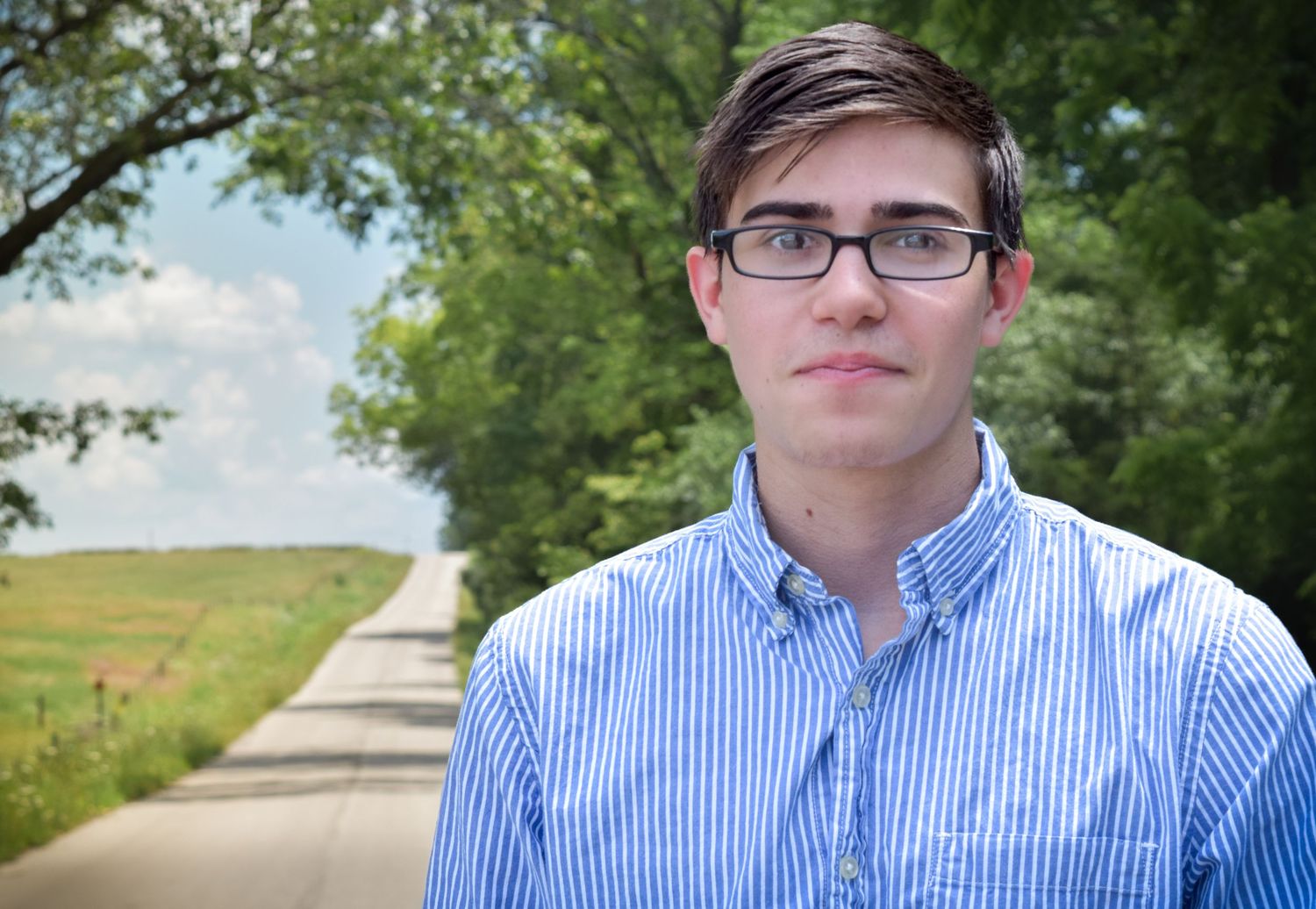 A person in a blue striped button-down shirt stands on a gravel road with green trees and fields in the background.