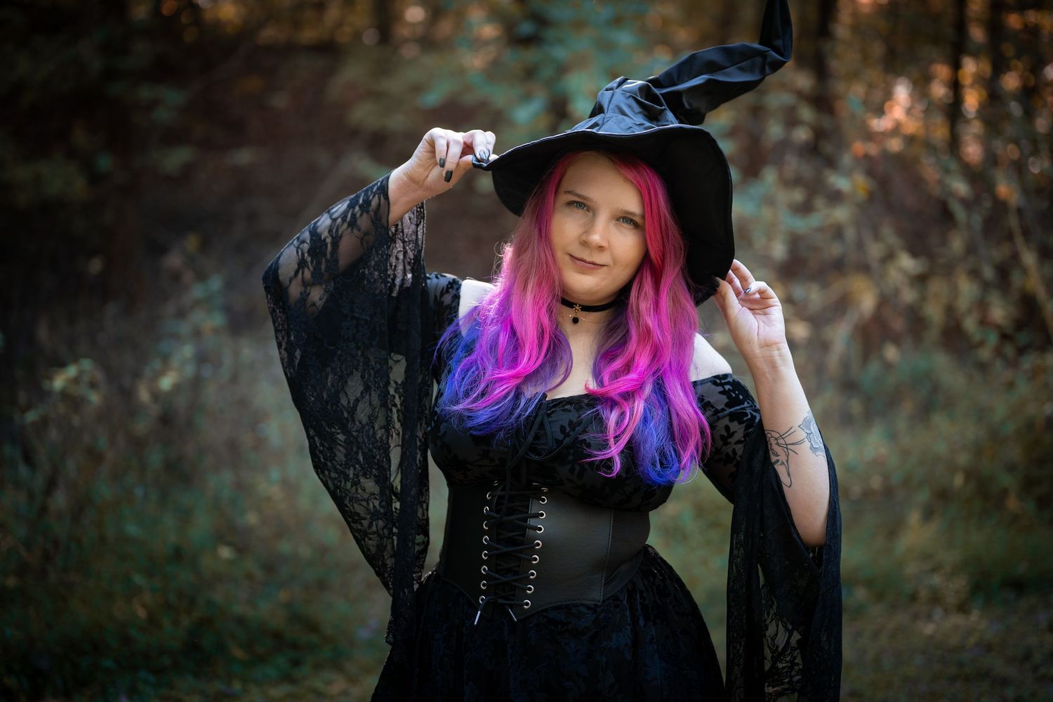 A person with vibrant pink and purple hair poses in a dark forest setting wearing a black lace outfit.