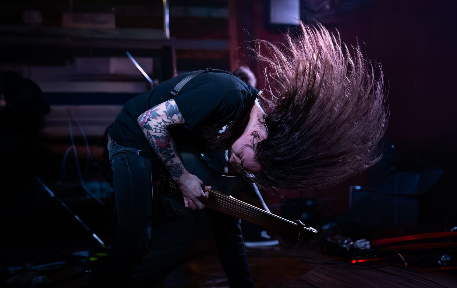 Tattooed rock musician headbanging on stage, hair flying, playing guitar under dramatic dark blue stage lighting.