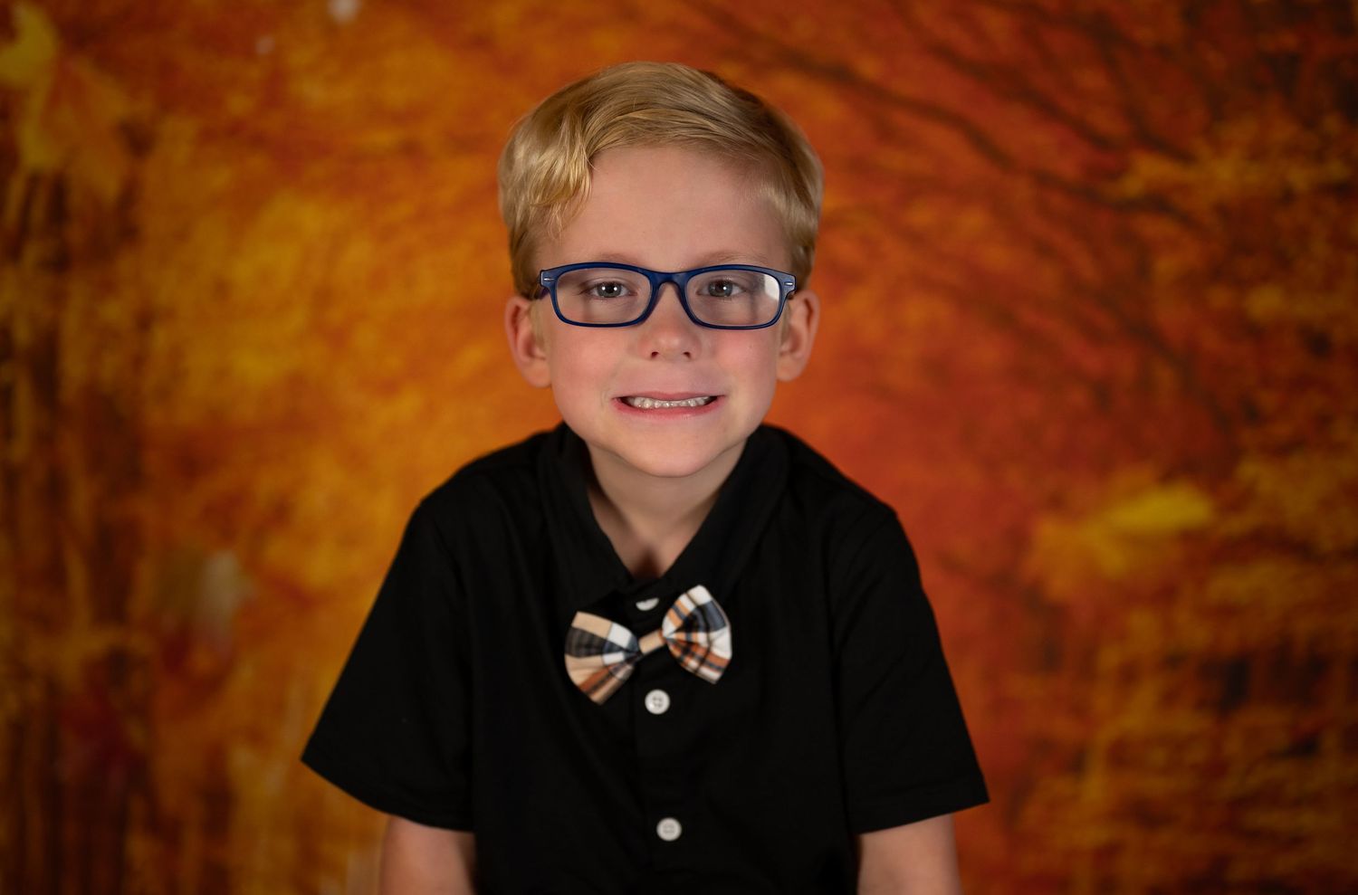 A child wearing glasses and a black dress shirt with bow tie smiles against an orange autumn background.