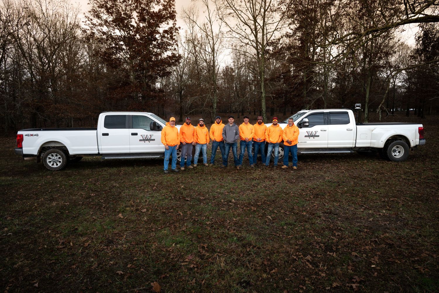 Line of workers in orange safety vests standing on hill with white trucks.