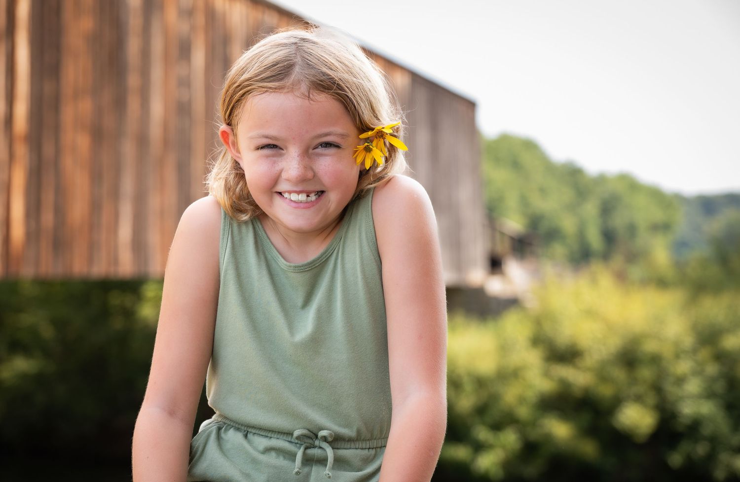 A child in a sage green tank top poses outdoors with a cheerful expression and yellow flower in their hair.