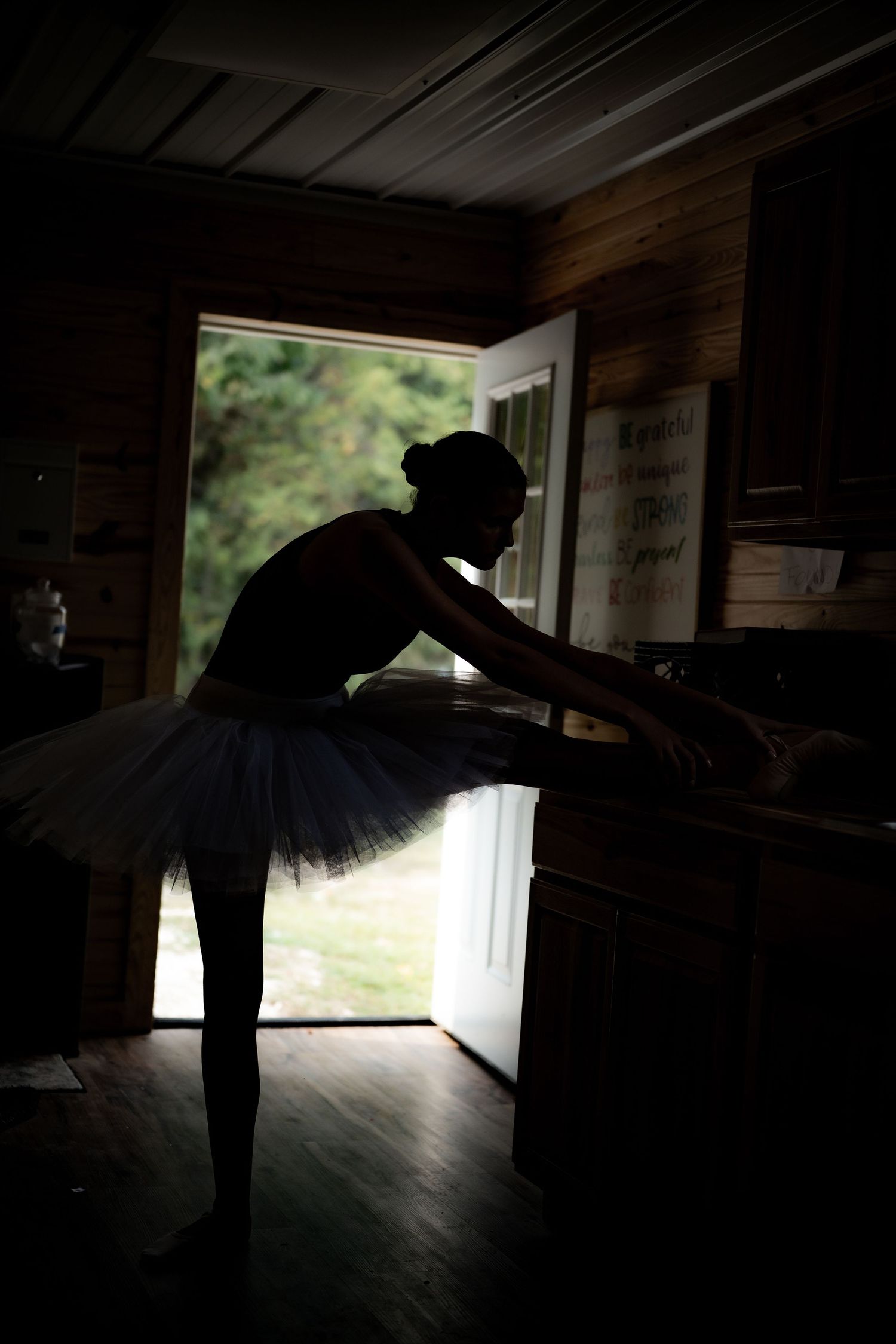 Silhouette of a ballet dancer in a tutu stretches at a barre against a bright doorway in a rustic wooden studio.