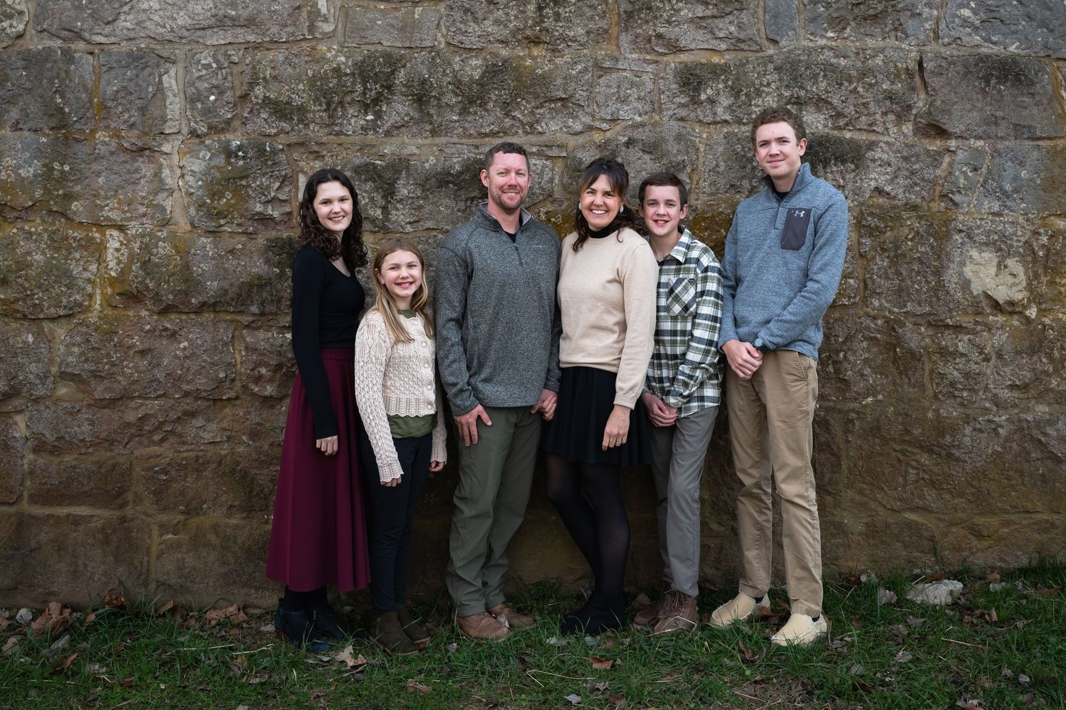 Family group poses for photo against weathered stone wall backdrop outdoors.