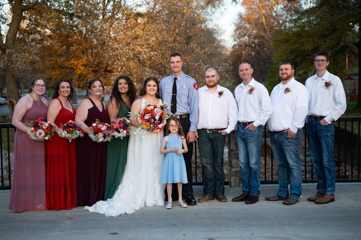 Large wedding party in burgundy and blue attire posed outdoors during autumn.