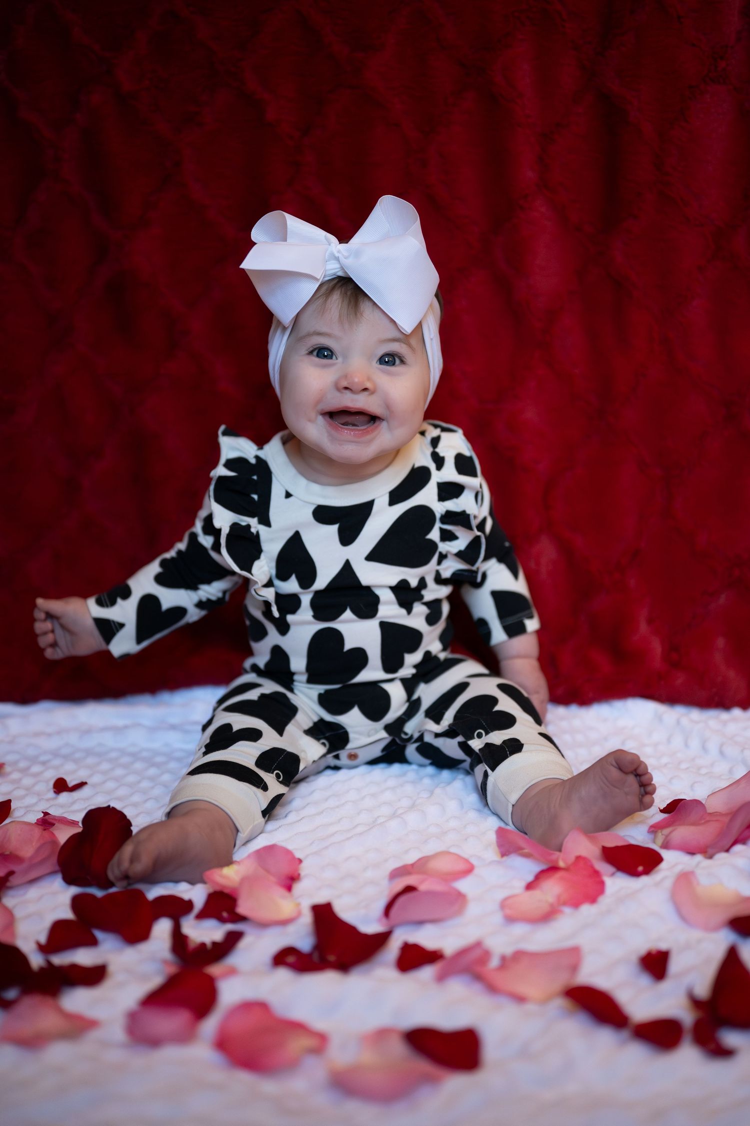 Baby in black and white heart print outfit lying on white blanket with scattered red and pink rose petals.