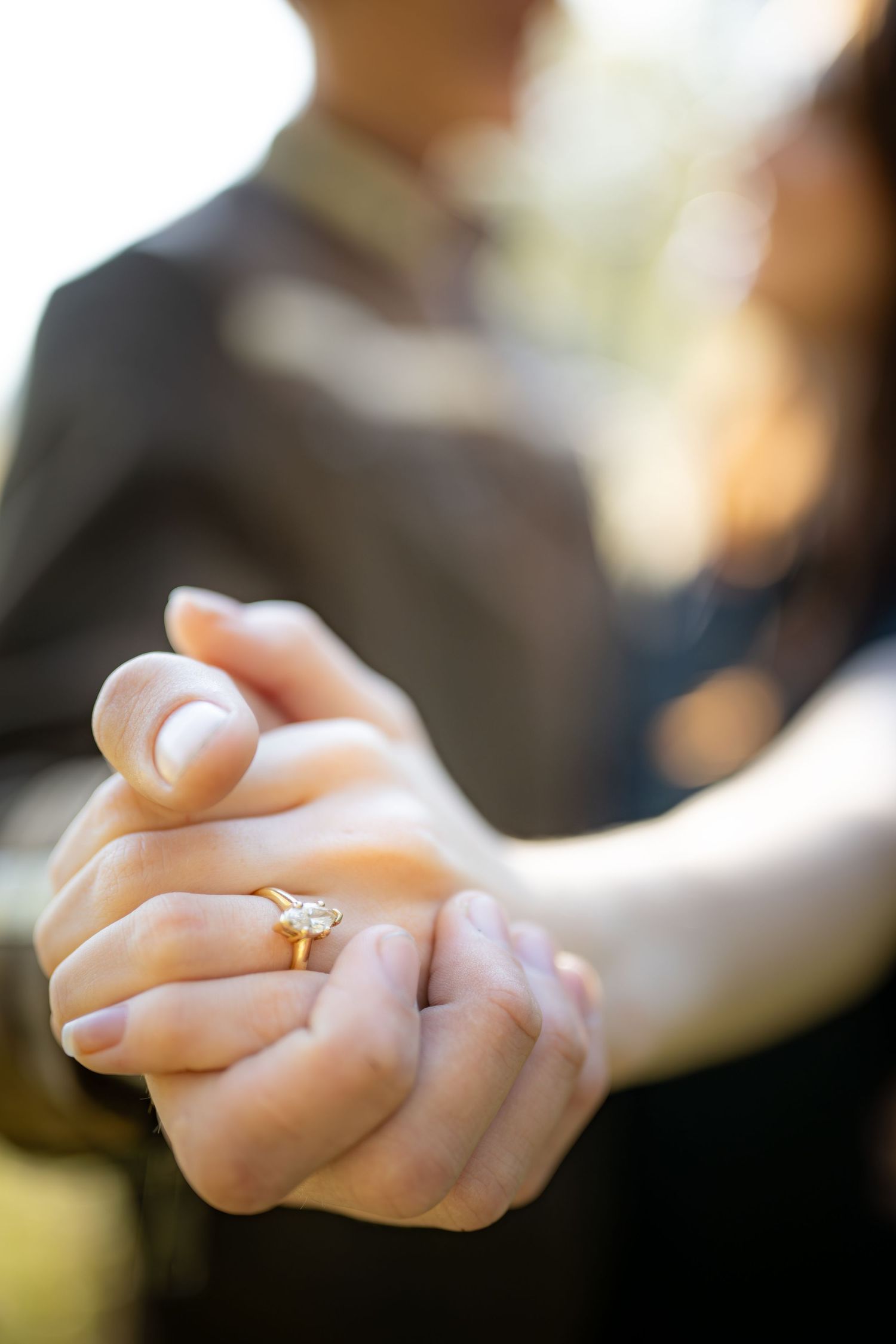 Two hands gently clasped together with a gold wedding ring visible in soft natural lighting.