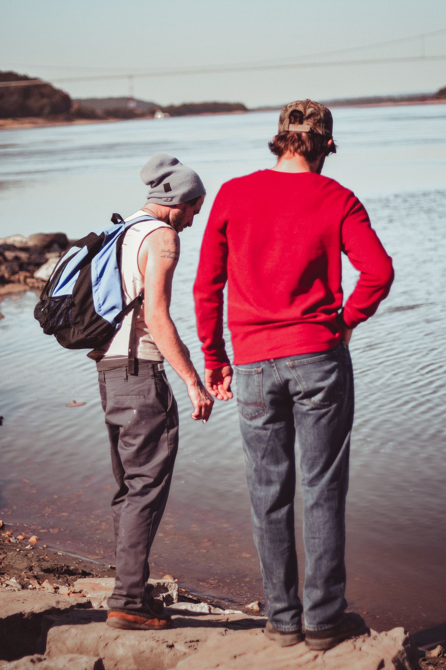 Two people in brown pants and casual wear stand at the edge of a lake looking at the water together.