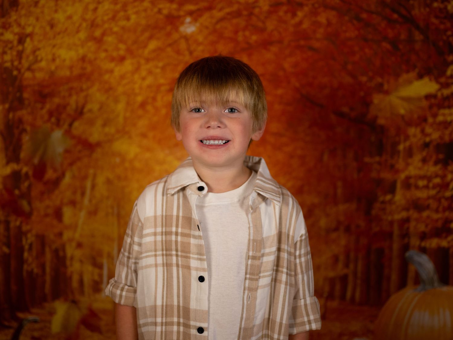 A child in a plaid button-down shirt smiles against a fall-themed backdrop with orange leaves.