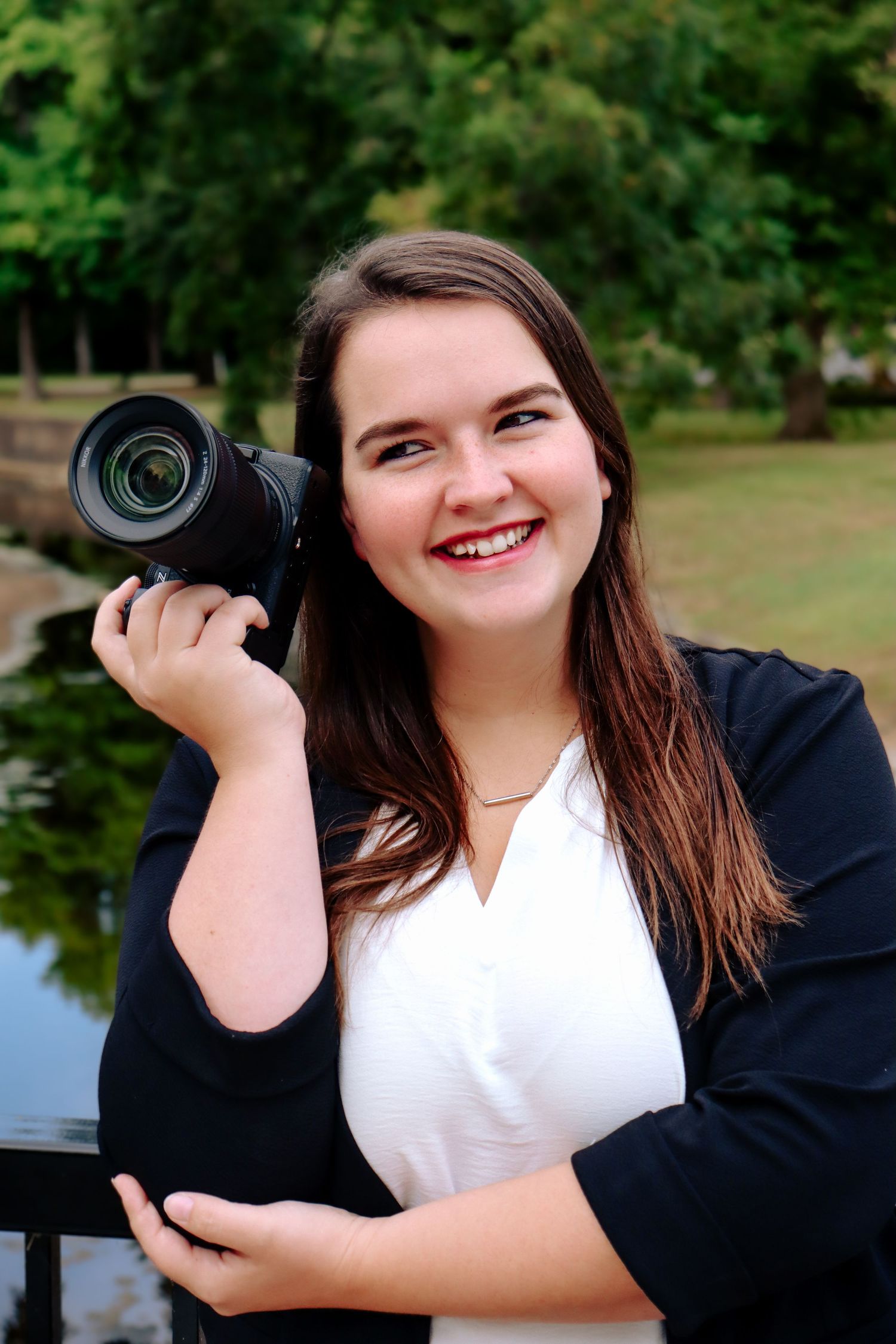 Professional photographer holding a DSLR camera with telephoto lens outdoors by a lake in a white and black outfit.
