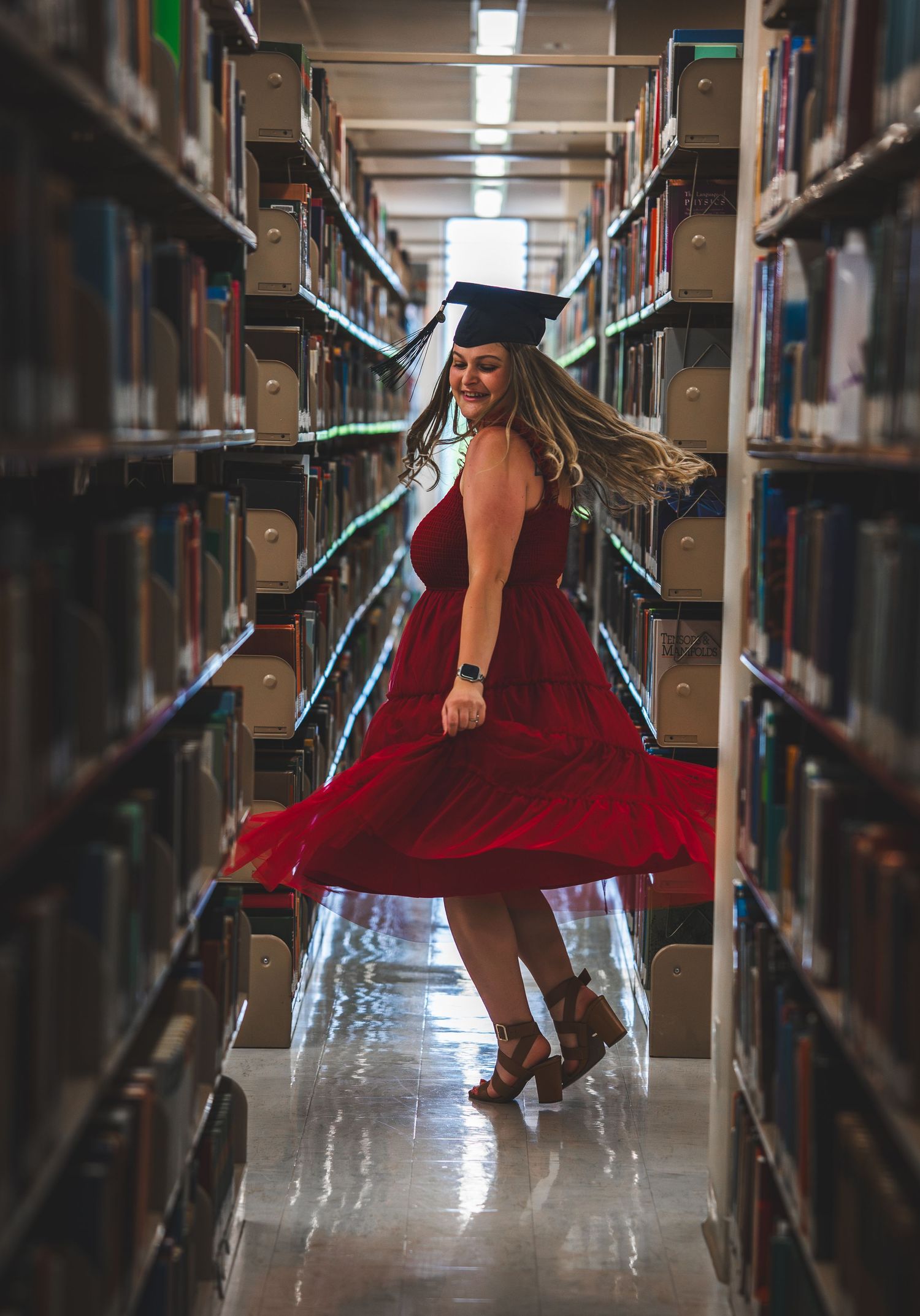 A woman in a red dress and graduation cap twirls joyfully between library bookshelves celebrating her academic achievement.