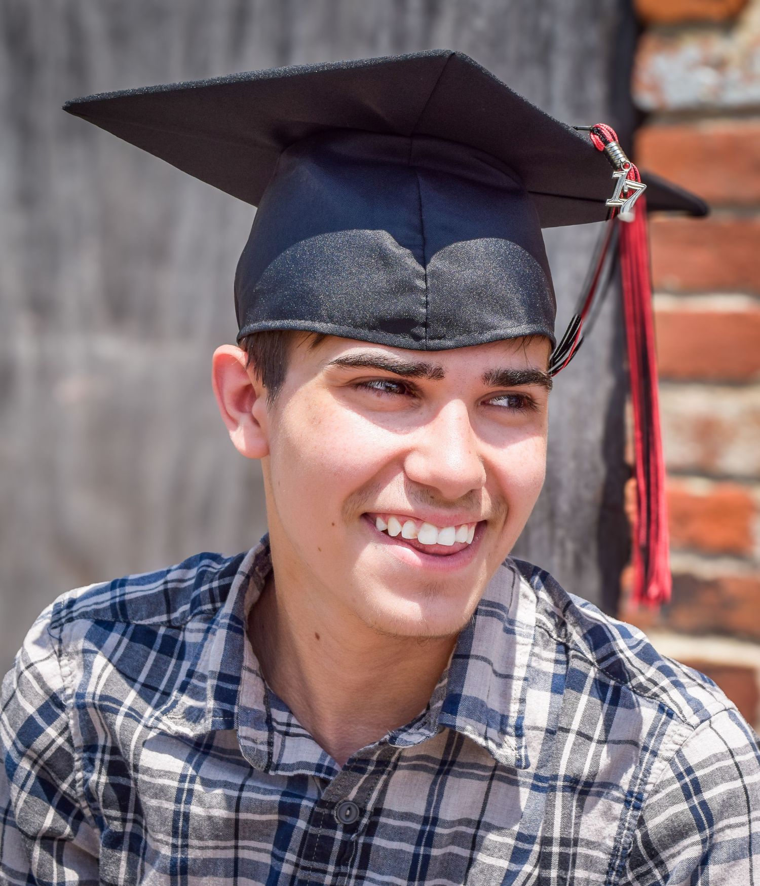 Graduate wearing plaid shirt and mortarboard cap smiles brightly during outdoor graduation photo session.