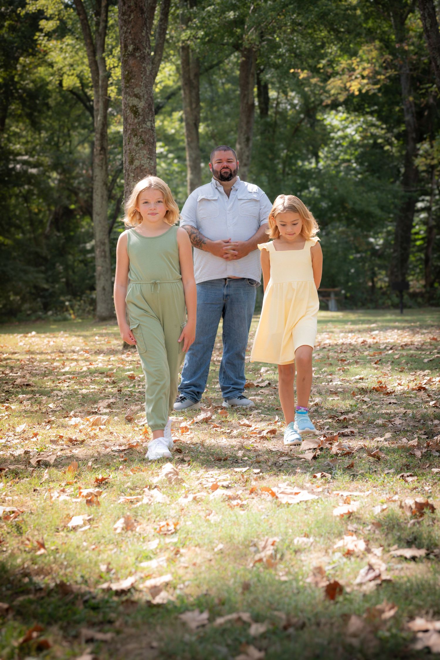 Three people stand together on fallen autumn leaves in a sunlit wooded area on a warm day.