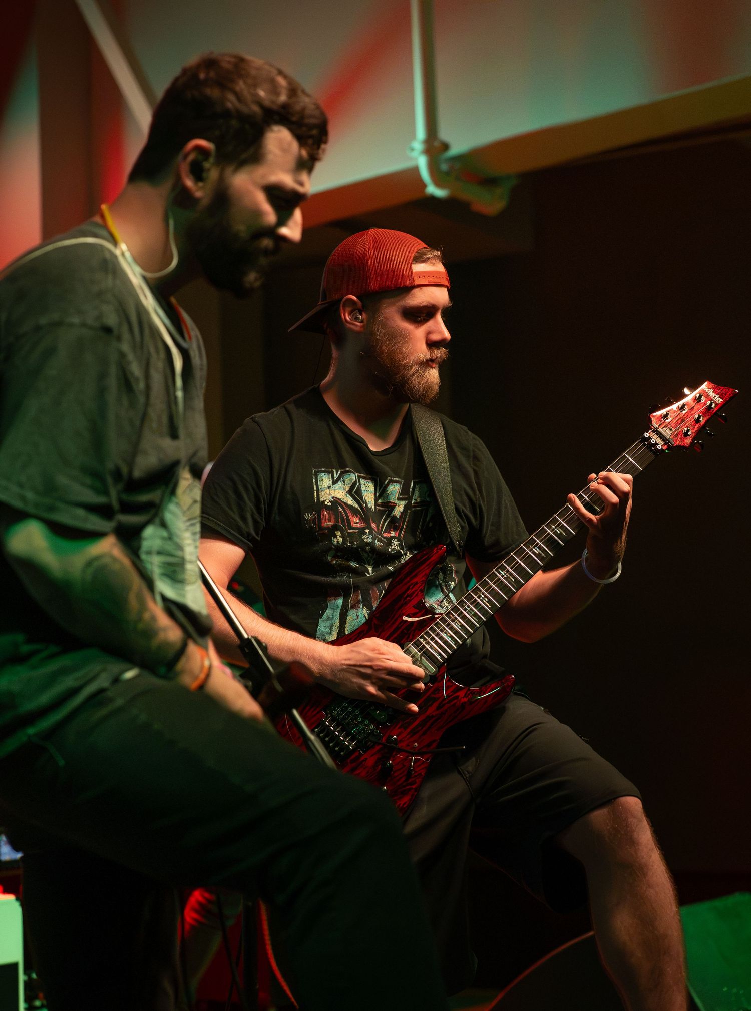 Two male guitarists playing electric guitars on stage, one wearing a red cap and band t-shirt, performing in moody stage lighting.