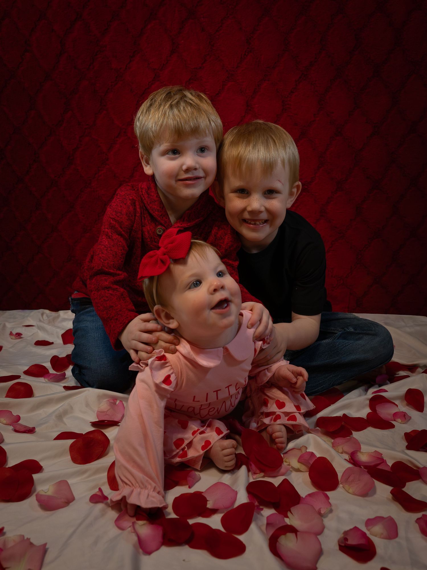 Children playing and laughing on white bedding scattered with red rose petals against a dark red backdrop.