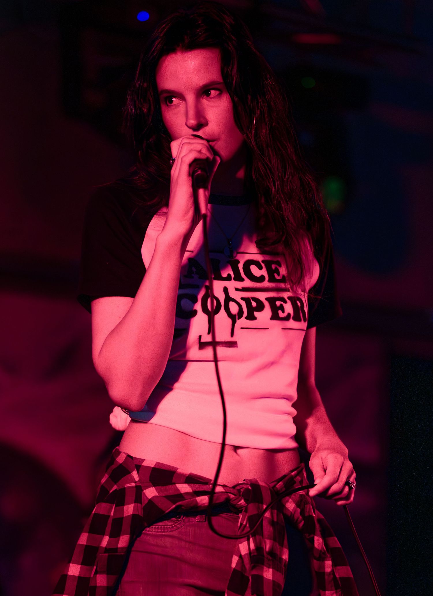 A performer in a white crop top and plaid skirt sings on stage under dramatic red lighting.