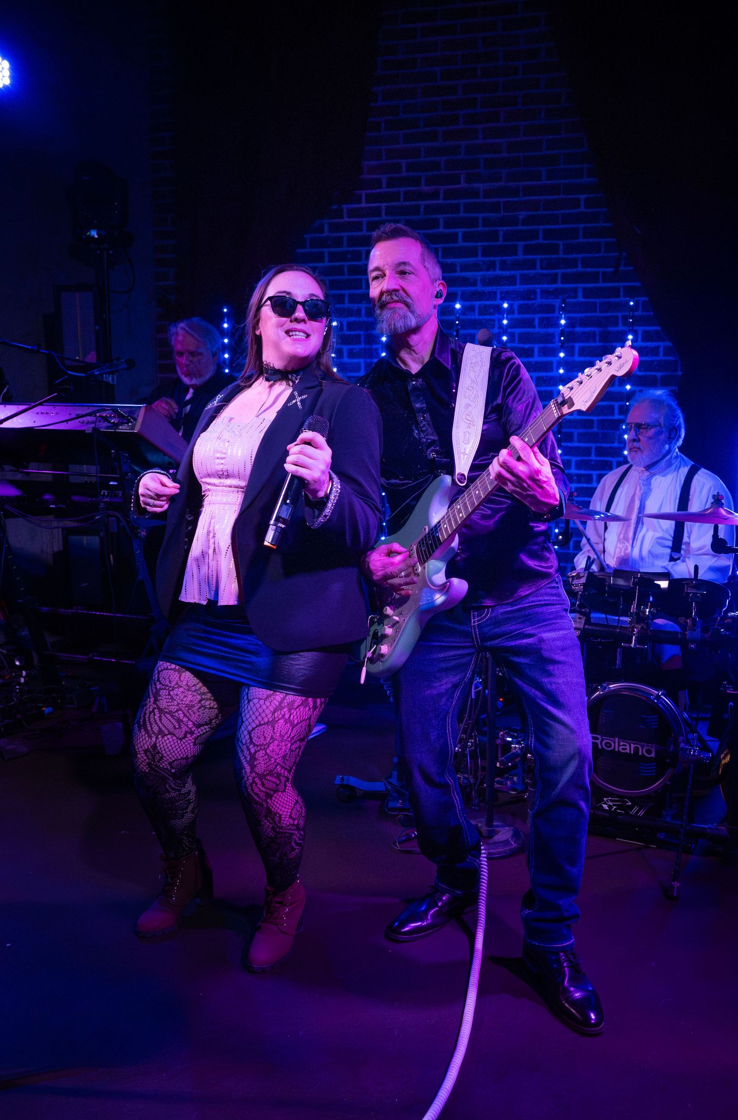 Musicians perform on stage in a dark venue with purple lighting while dancers move to the music on the dance floor.