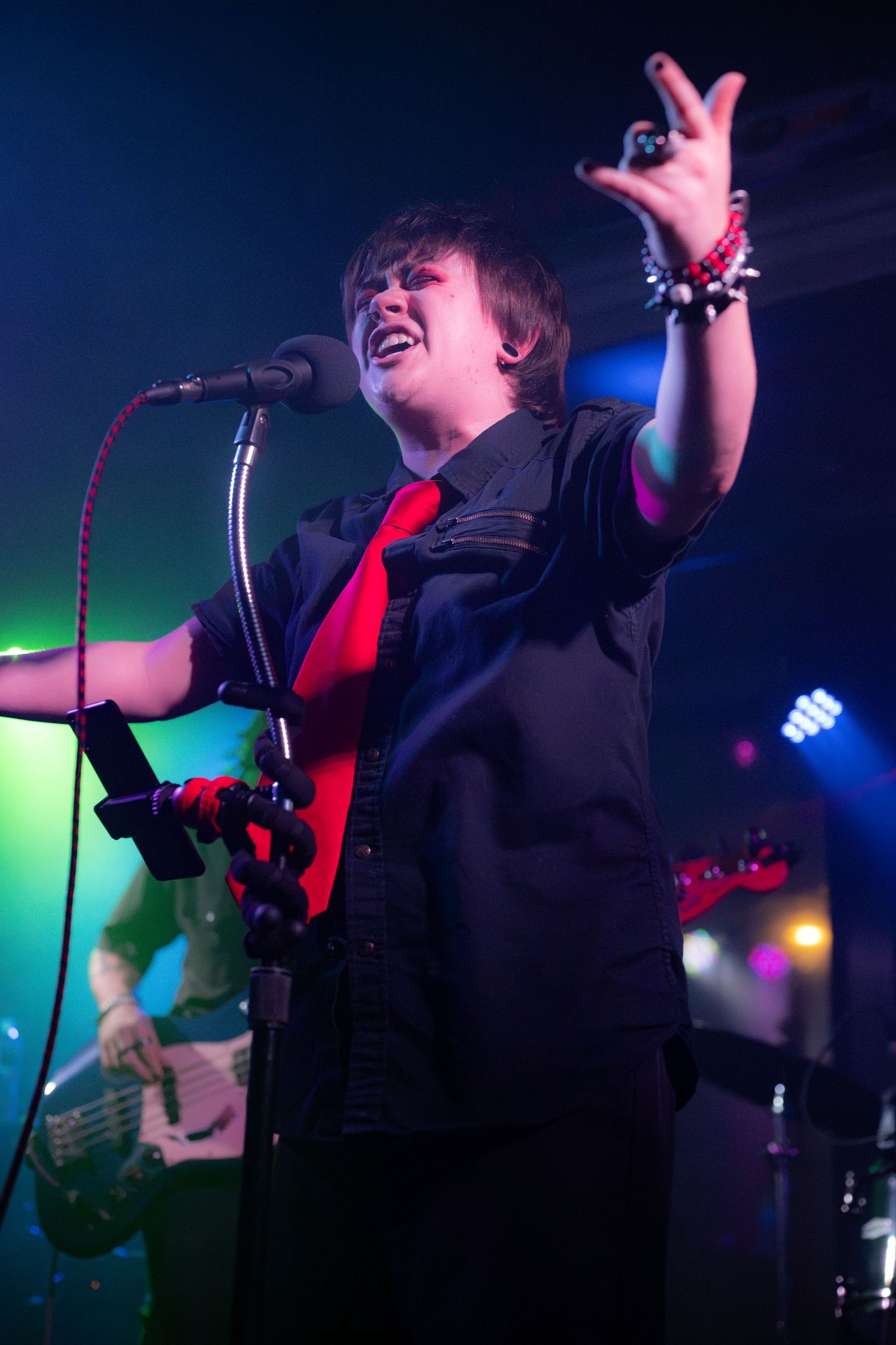 Young rock singer in black shirt and red tie performs passionately on stage with band members visible behind.