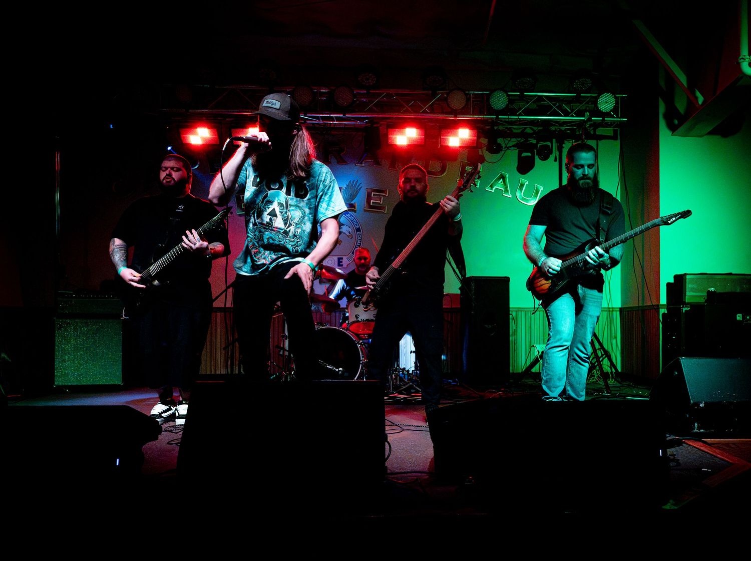 Rock band performing on a dark stage with green and red lights, featuring guitarist, vocalist, bassist, and drummer.