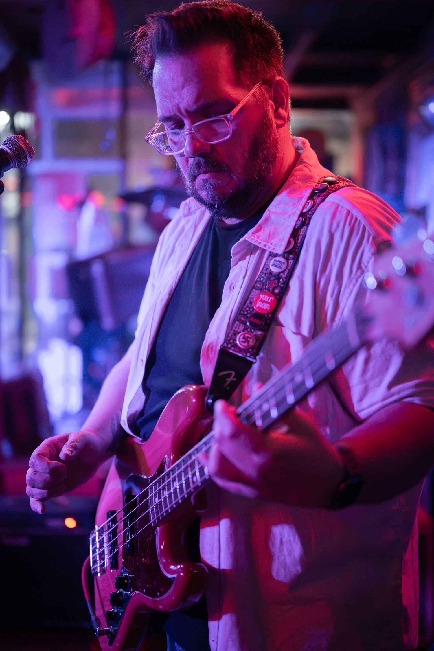 Middle-aged man with glasses playing red electric guitar on stage under purple and pink lights at a live music venue.