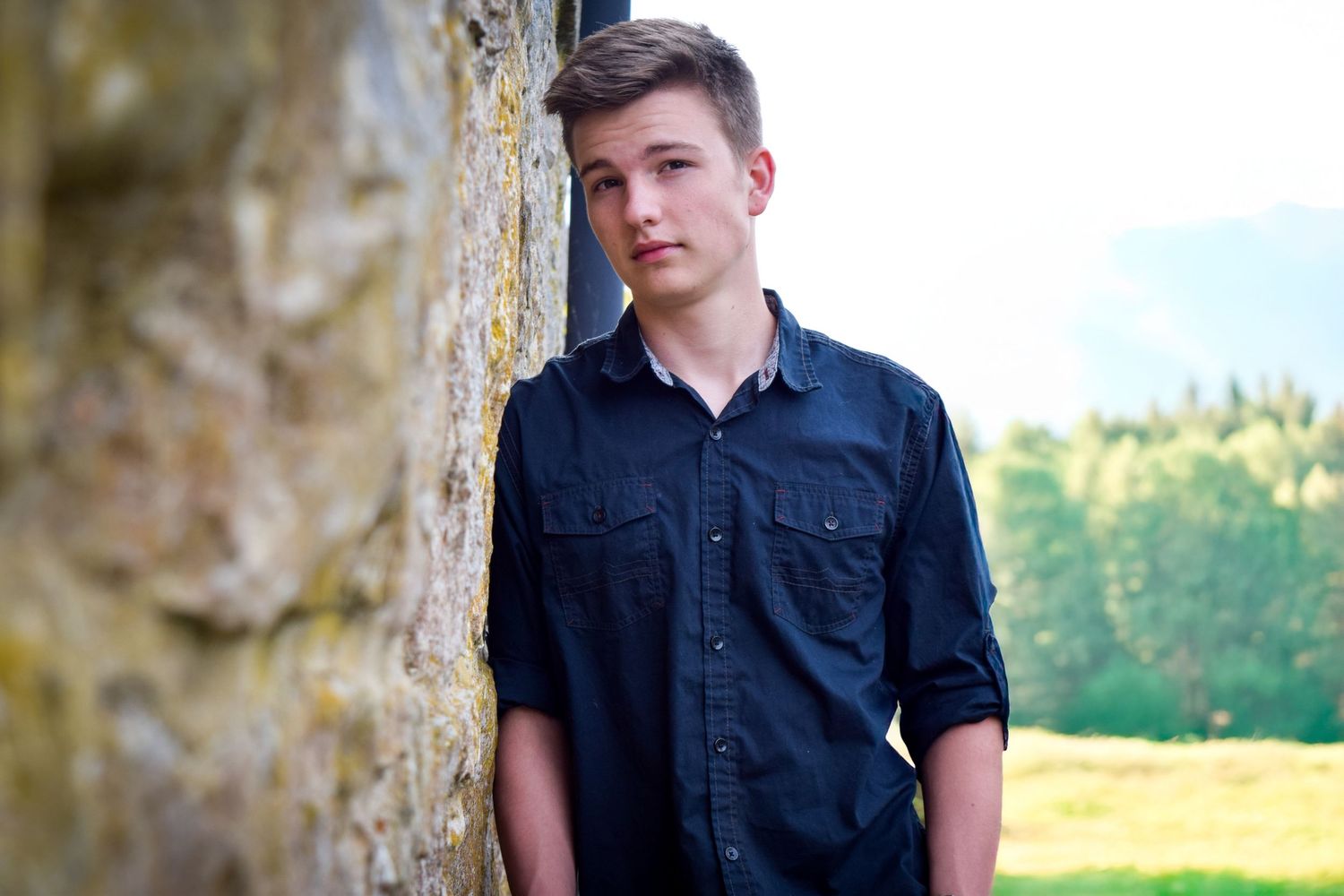 A person in a navy blue button-down shirt poses outdoors against a stone wall with trees and fields in the background.