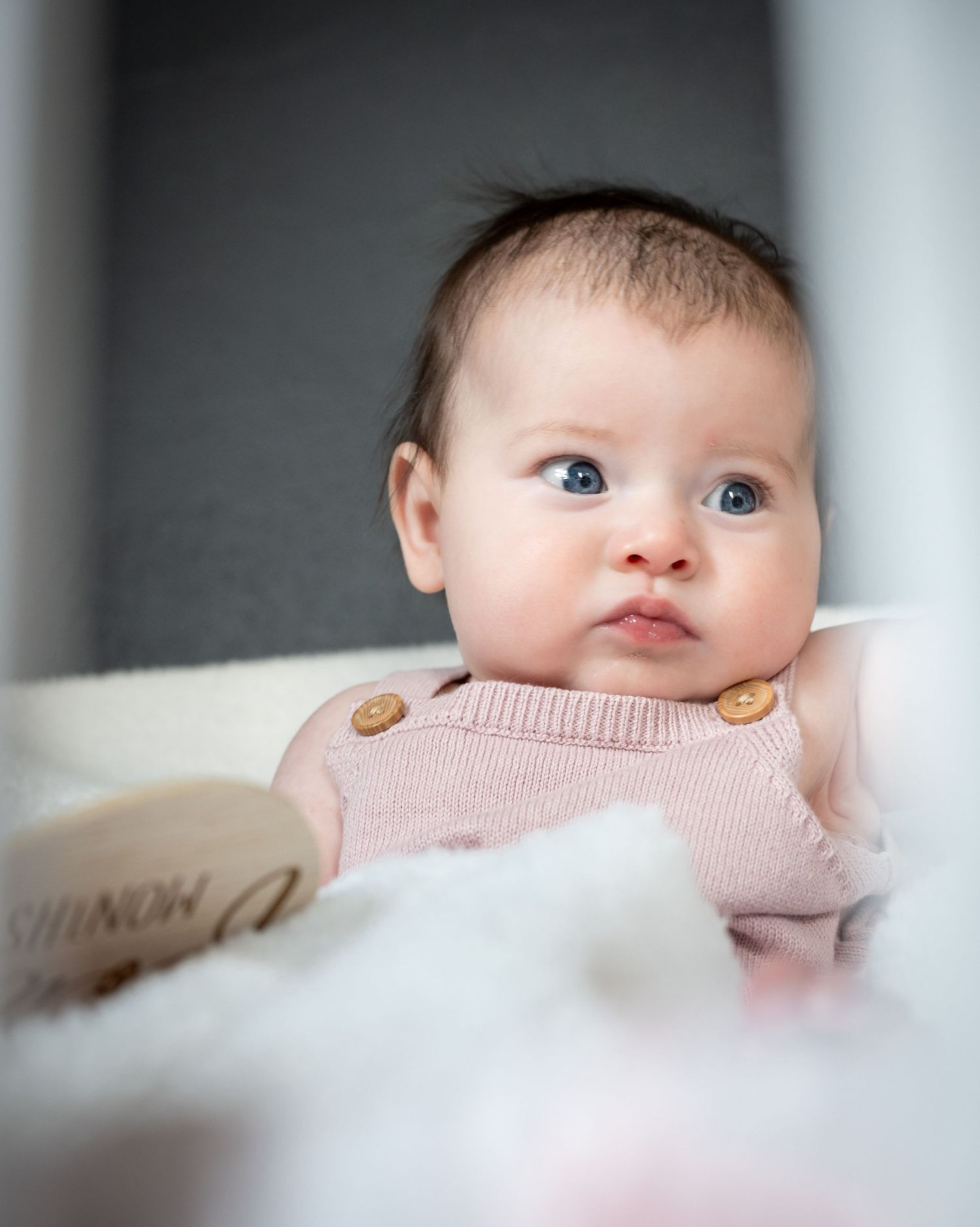 A baby wearing a soft pink knitted sweater with button details gazes upward with bright blue eyes.