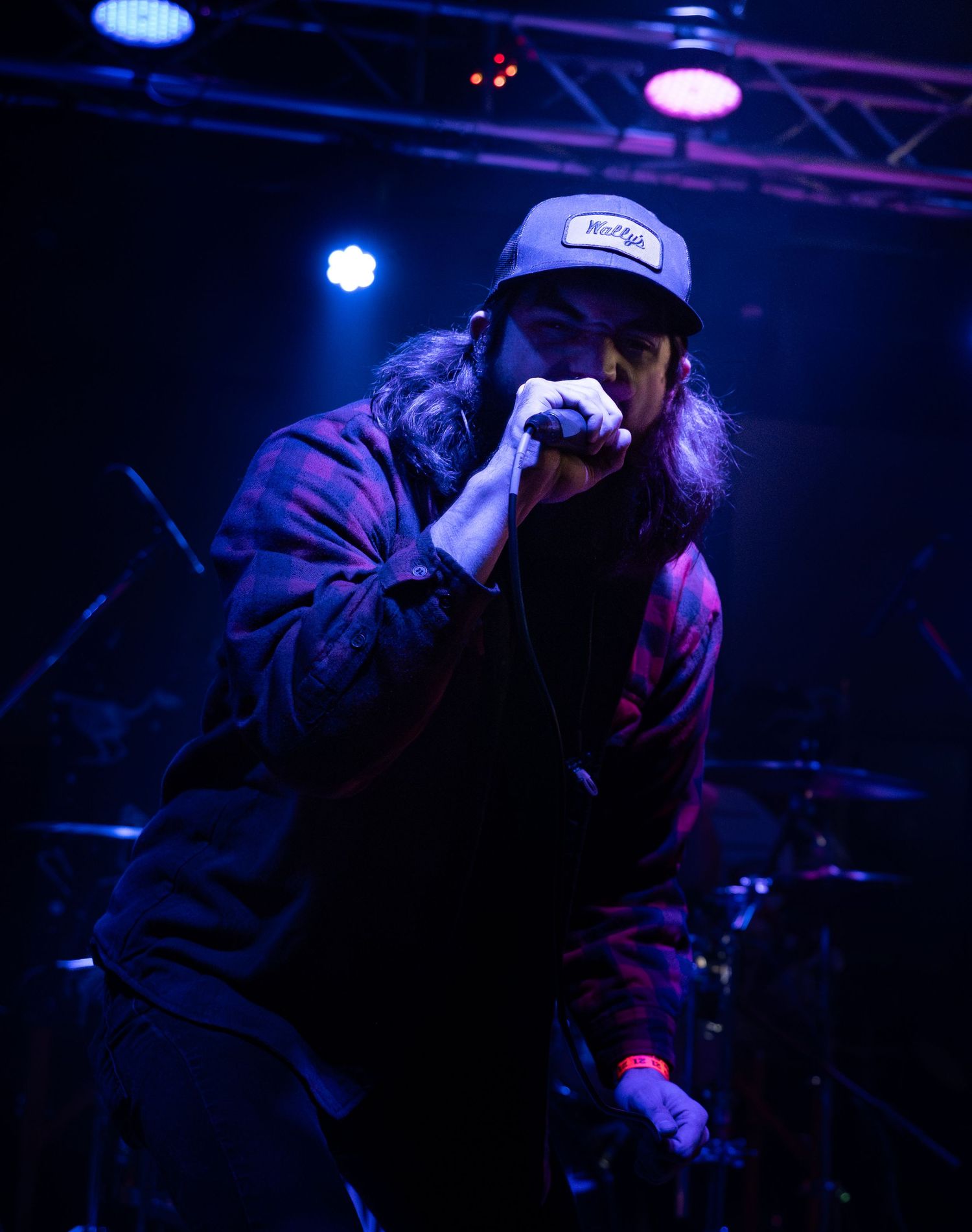 A performer in dark lighting holds a microphone while wearing a baseball cap during a nightclub performance.