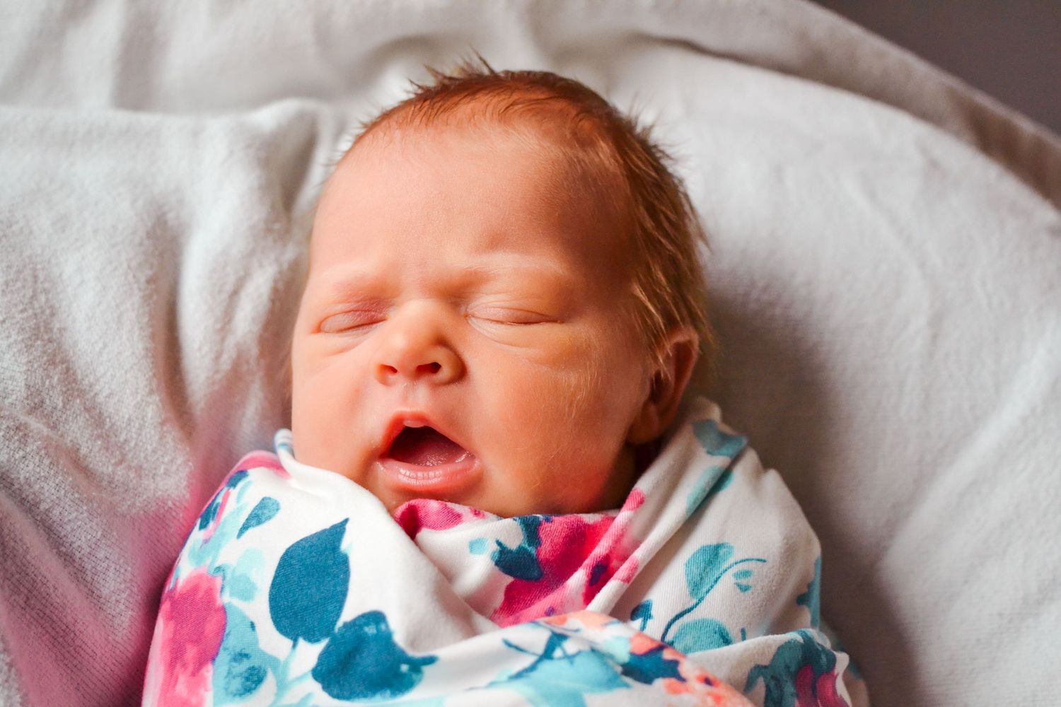 A sleeping newborn baby wrapped in a white and turquoise floral swaddle blanket rests peacefully on white bedding.