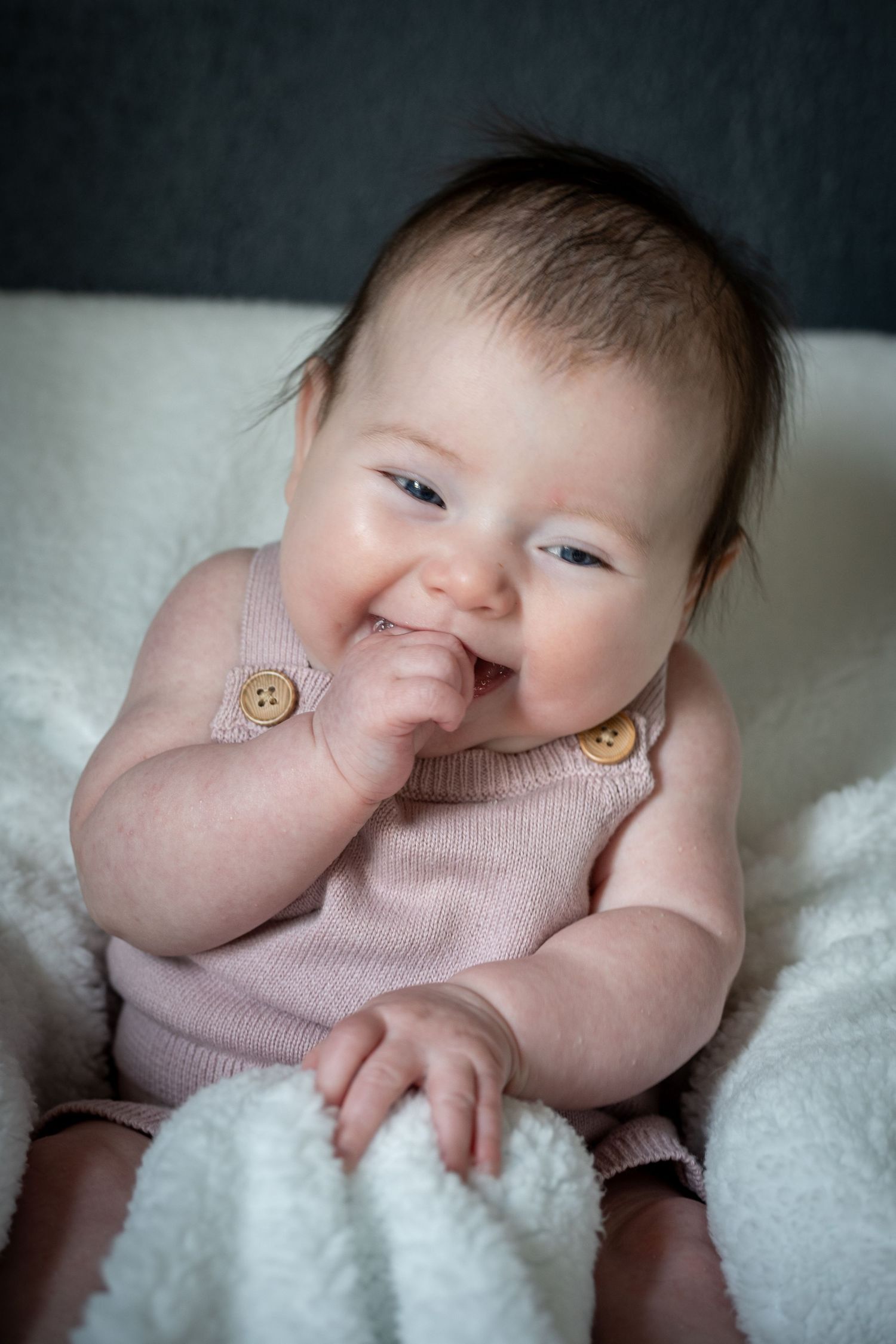 A smiling baby wearing a pink knit romper with wooden buttons lies on a white blanket.