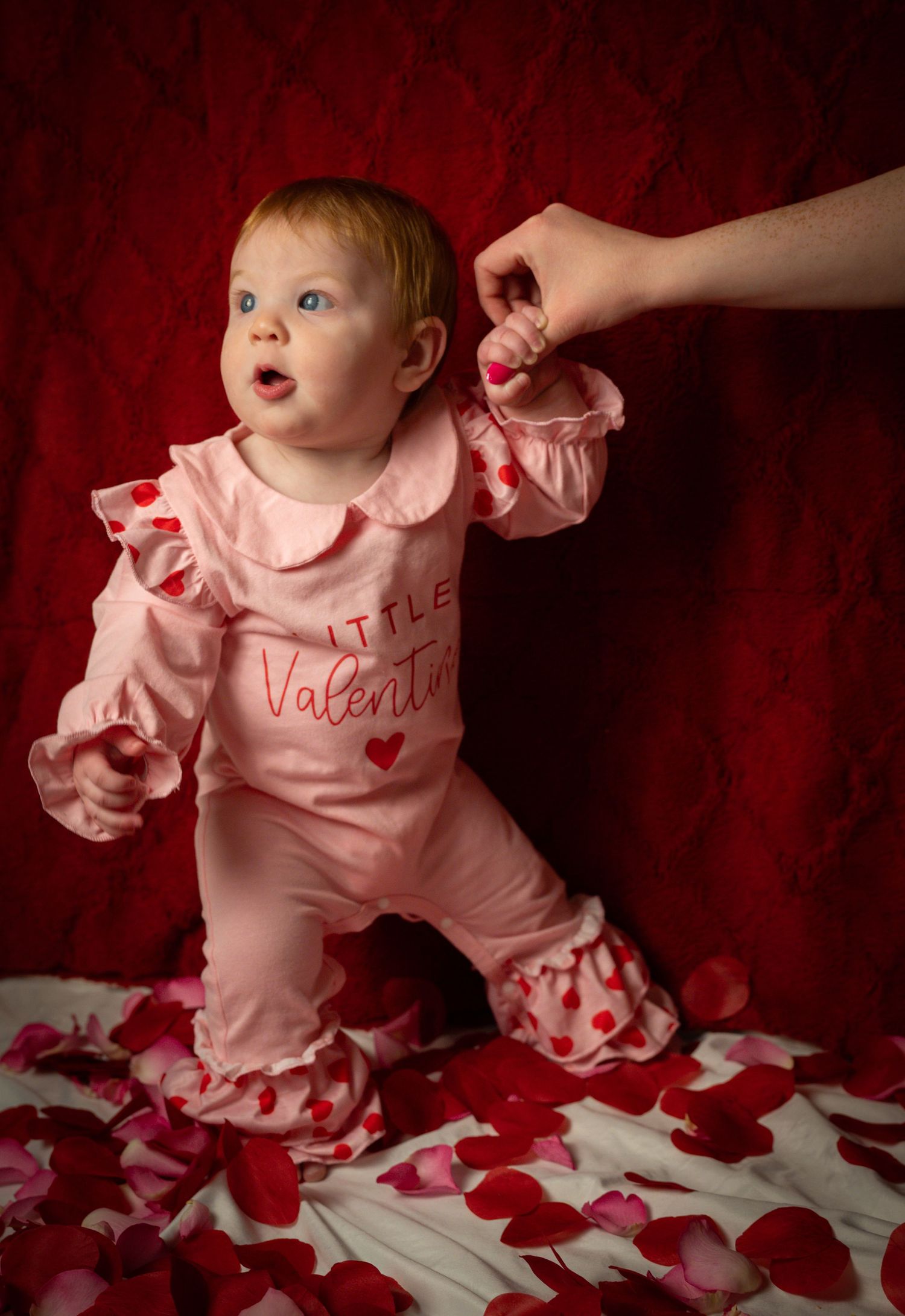 Baby in pink ruffled Valentine's outfit sits among scattered red rose petals against dark red backdrop.