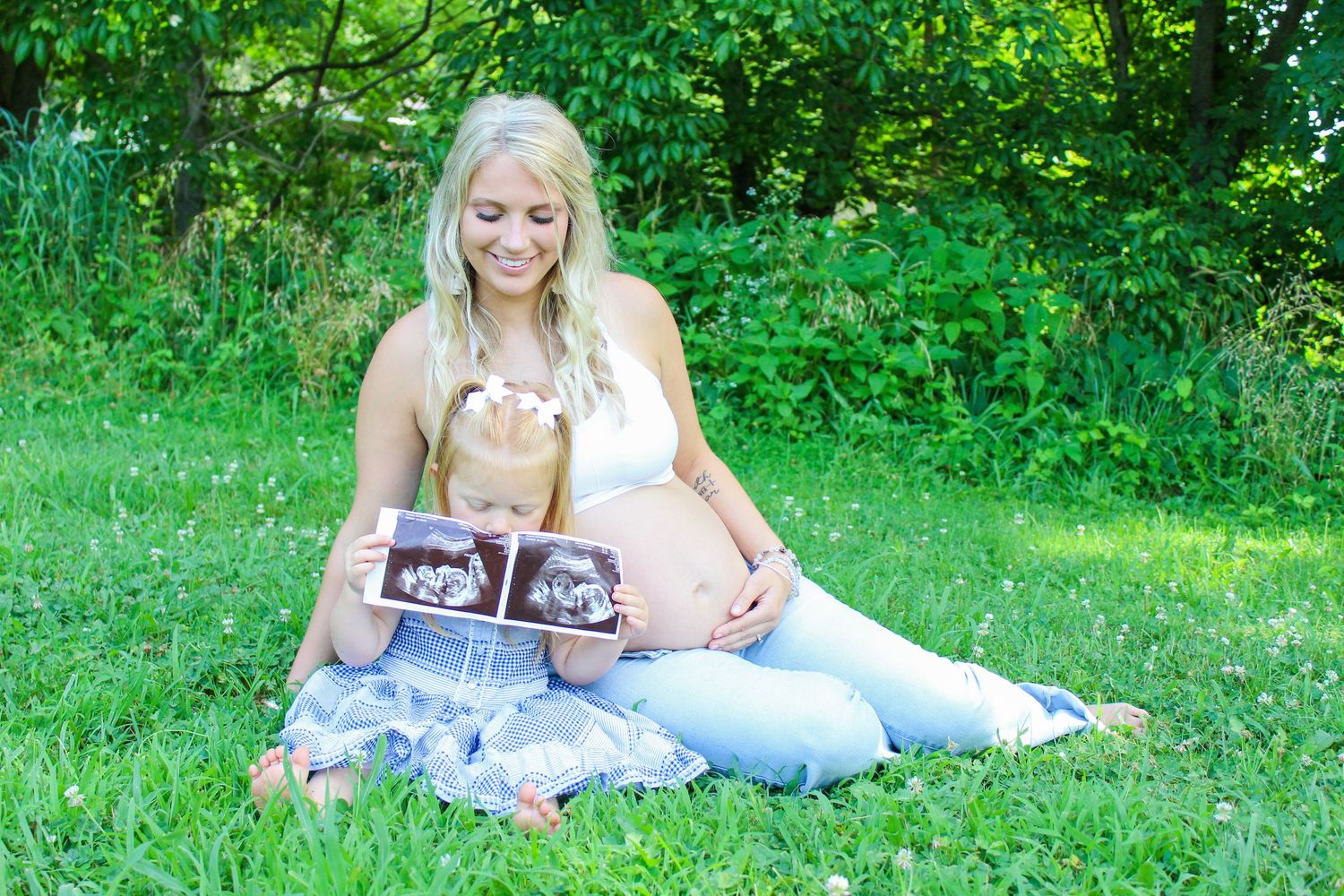 A pregnant woman sits on grass holding ultrasound photos during an outdoor maternity session.