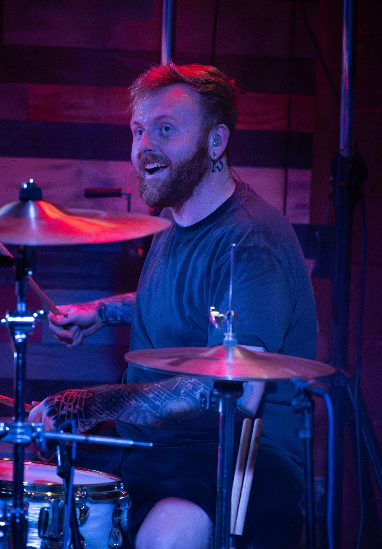 Tattooed male drummer with red beard performing on stage under colorful purple and red lighting at a live music venue.