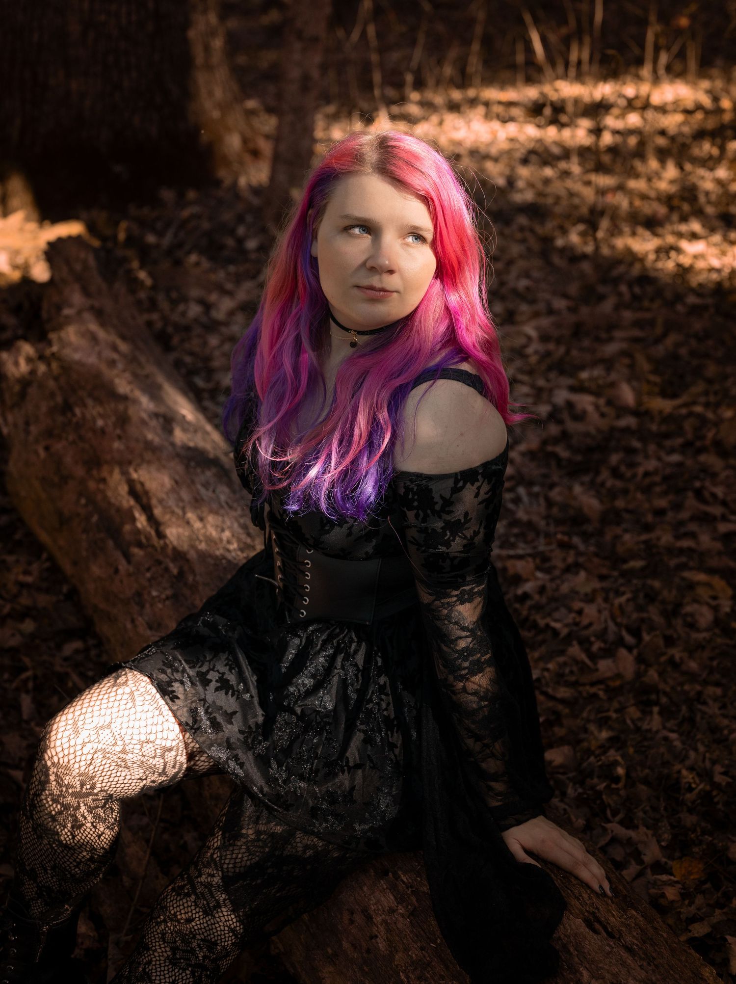 A person with vibrant pink and purple hair poses in a black lace dress against a moody forest backdrop in golden light.